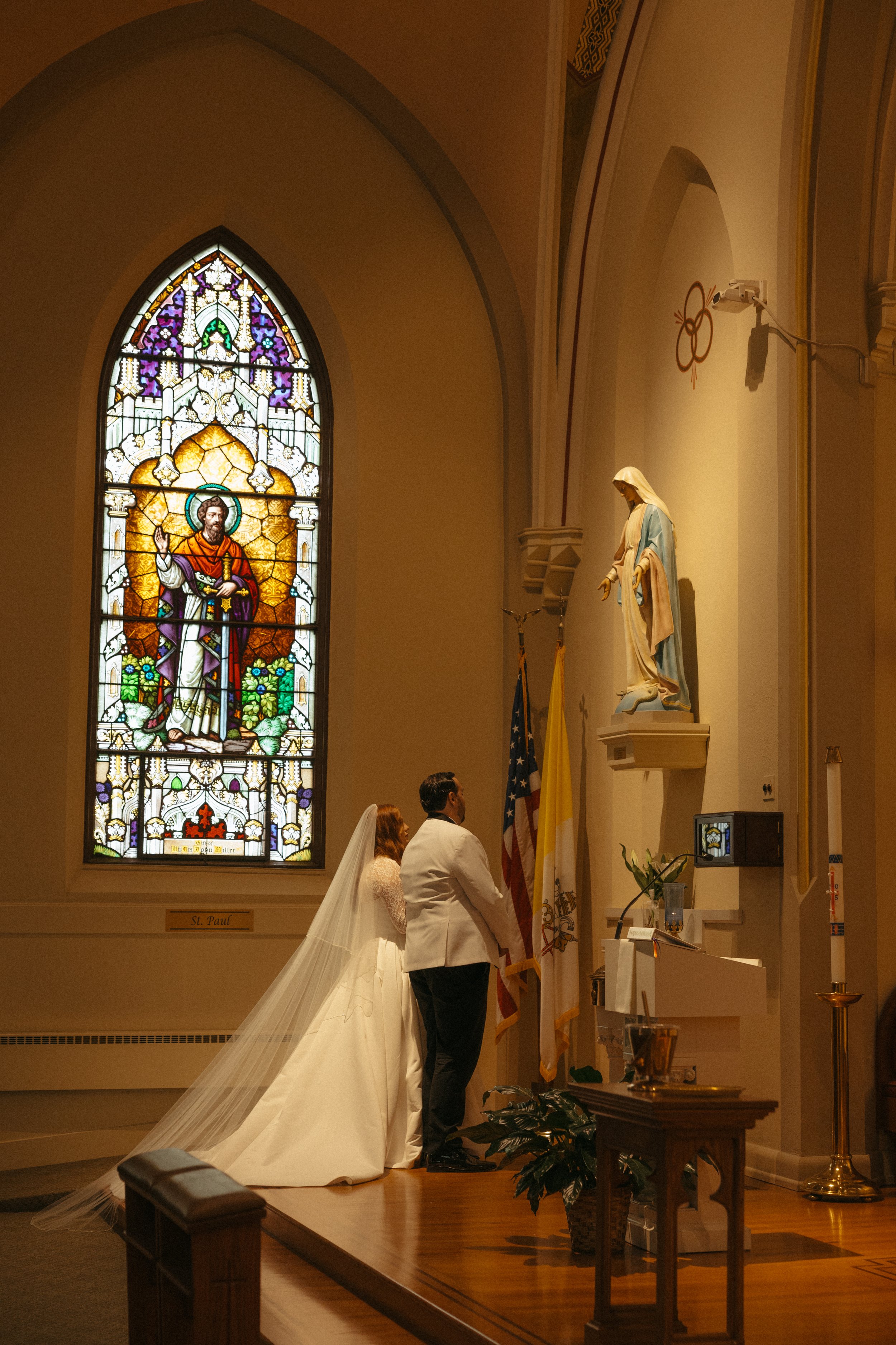 Isabel Marie Photography - A bride and groom standing at the altar in a church, with stained glass windows of Jesus and a statue of the Virgin Mary.