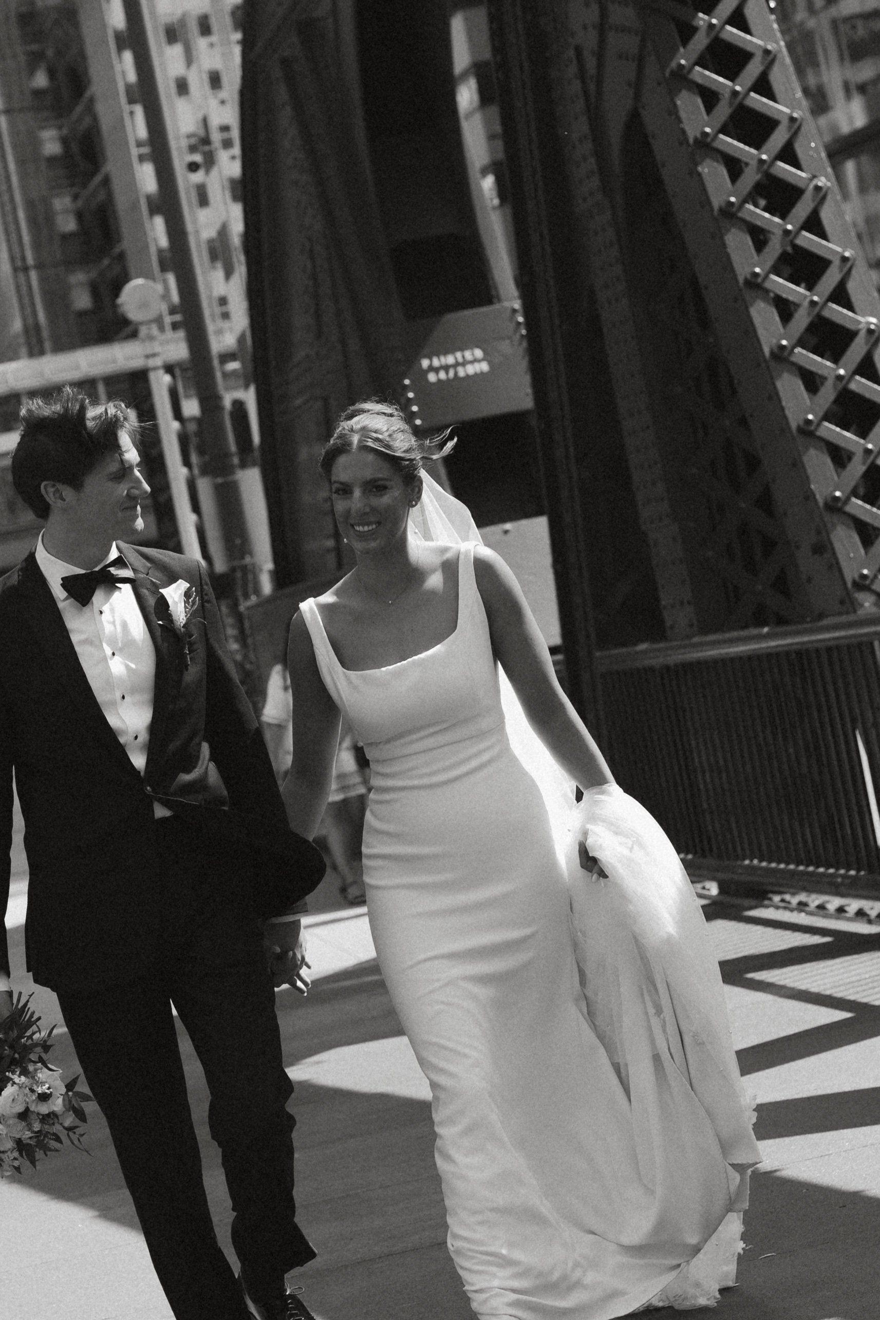 Isabel Marie Photography - A black and white photo of a bride and groom walking hand-in-hand outdoors in an urban setting, a woman smiling and holding her wedding dress.