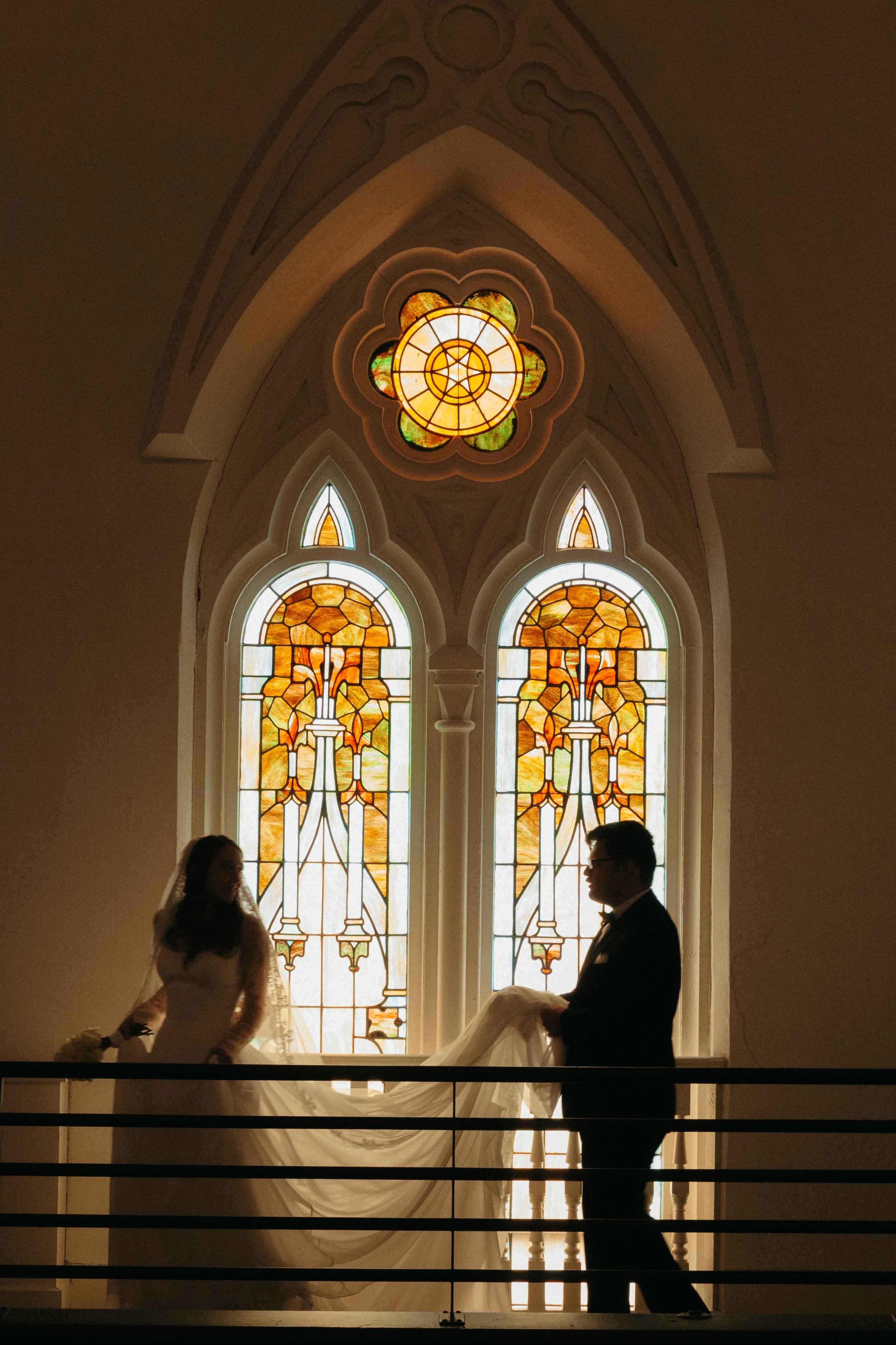 Isabel Marie Photography - Silhouettes of a bride and groom in front of stained glass windows in a church, with the groom holding the bride's veil or train.
