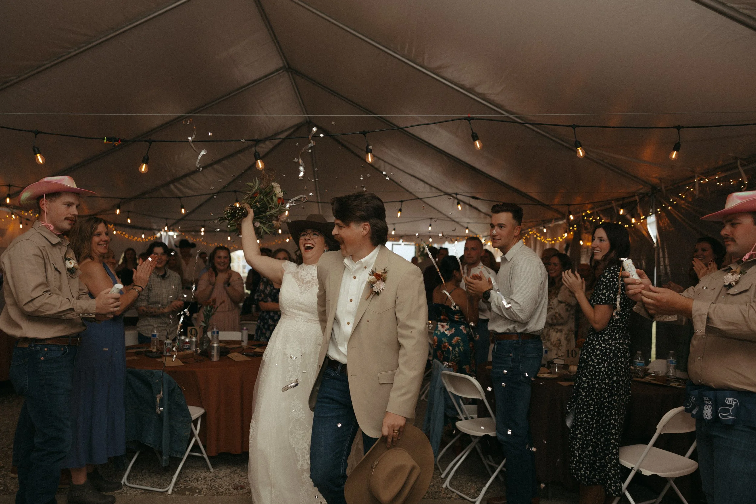 Bride and groom celebrating with wedding guests under a decorated tent, with string lights overhead. The bride is wearing a white dress and holding a bouquet, while the groom is in a beige jacket and holding a hat. Guests are clapping and smiling, dr