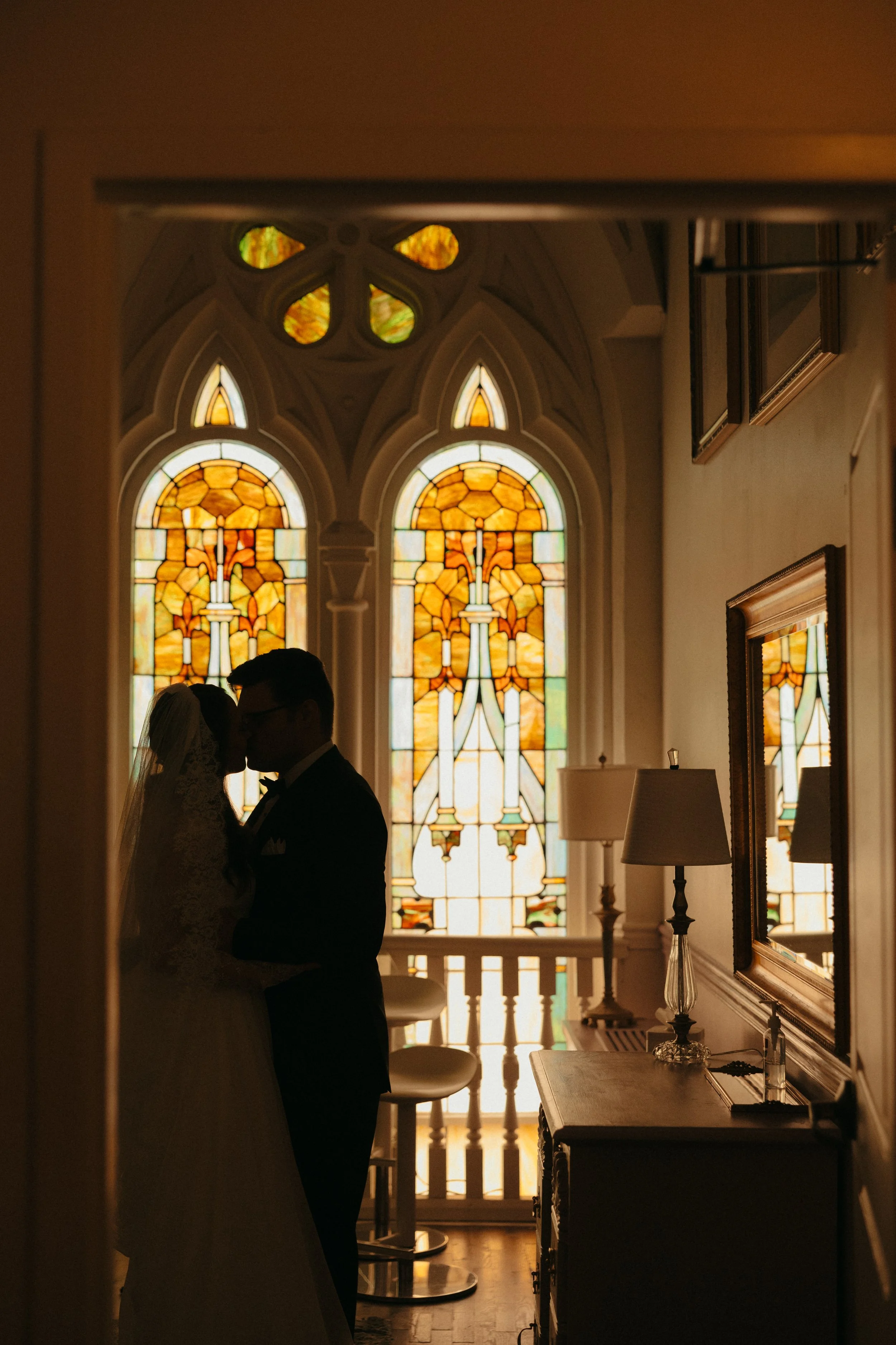 Isabel Marie Photography - Silhouetted bride and groom kissing in front of stained glass windows inside a church or chapel.