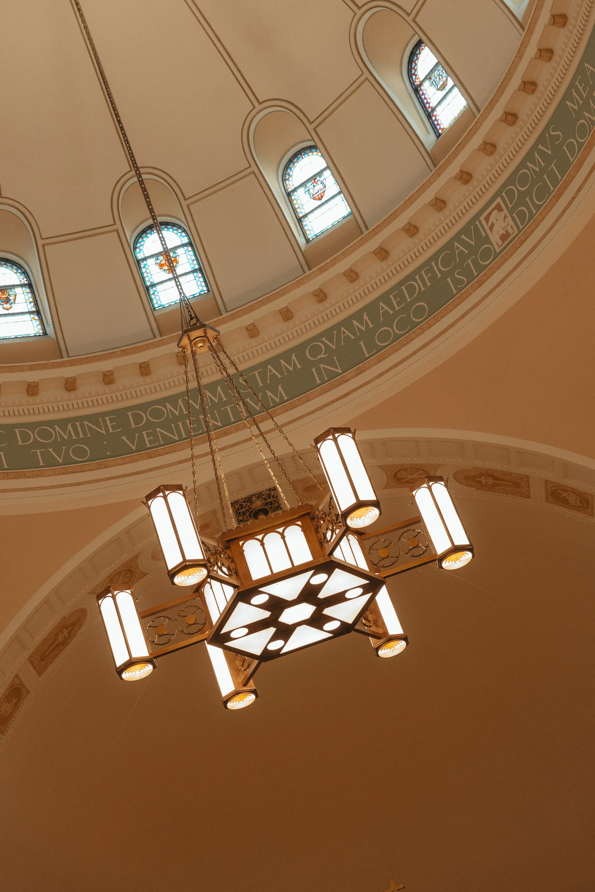 Isabel Marie Photography - Interior ceiling view of a church or cathedral showcasing stained glass windows, an ornate chandelier hanging from the ceiling, and Latin inscription around the dome.