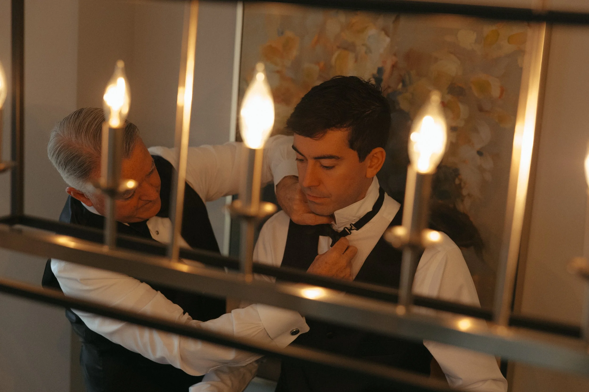 Isabel Marie Photography - An older man helping a young man with his bow tie in a dimly lit room, seen through a decorative metal gate with candle-like lights in the foreground.
