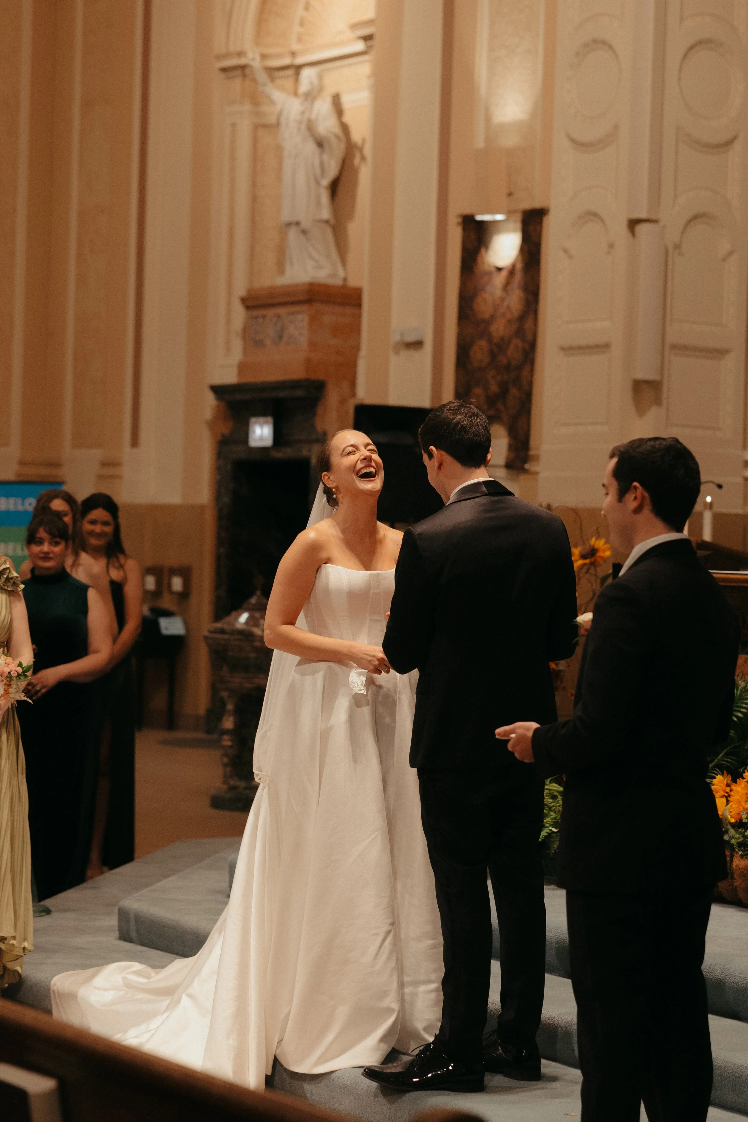 Isabel Marie Photography - A bride and groom exchanging vows and laughing during a wedding ceremony in a church, with guests watching in the background.