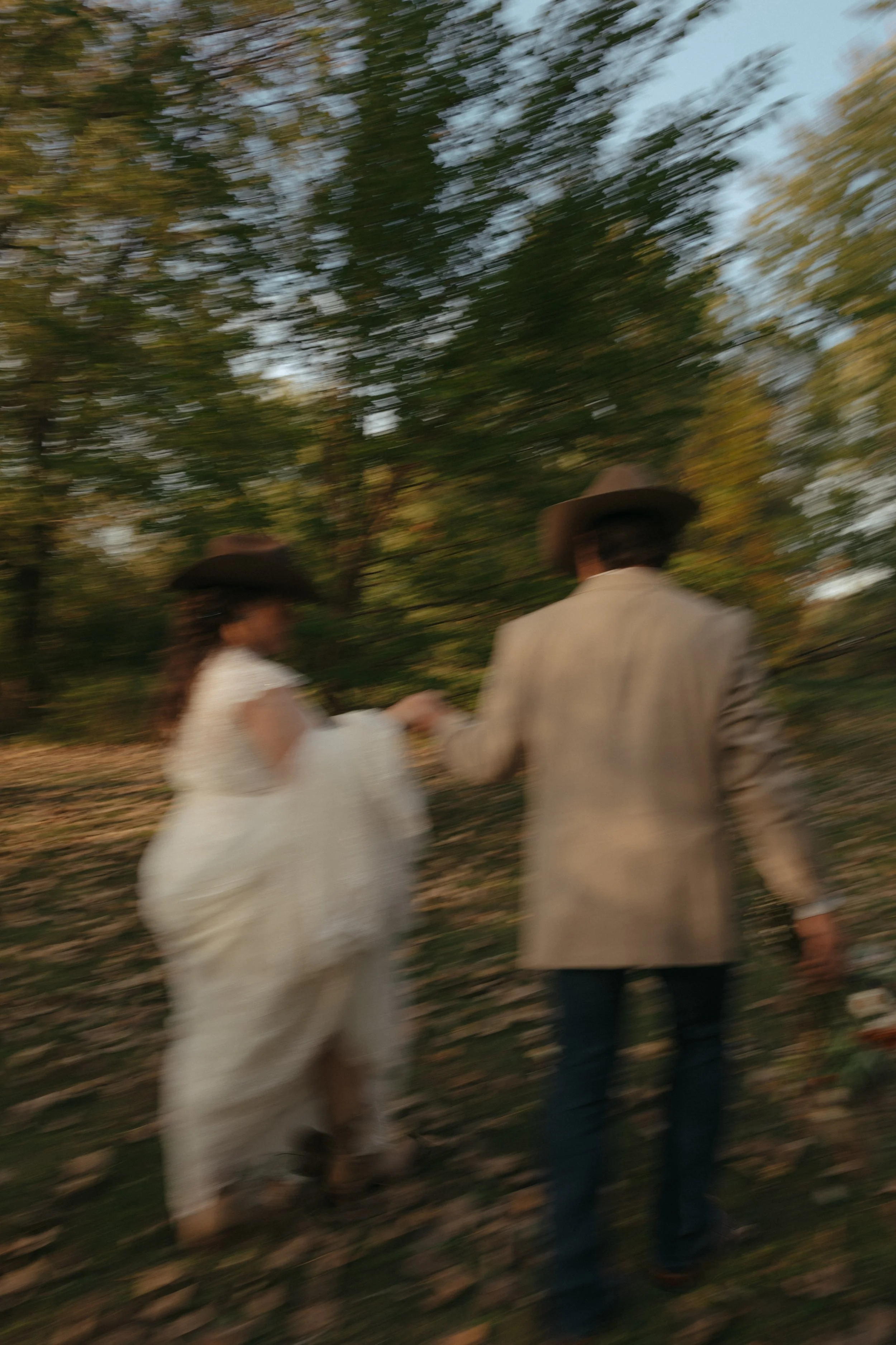 Isabel Marie Photography - Blurred image of a couple holding hands walking outdoors through a wooded area during daytime, wearing hats and casual attire.