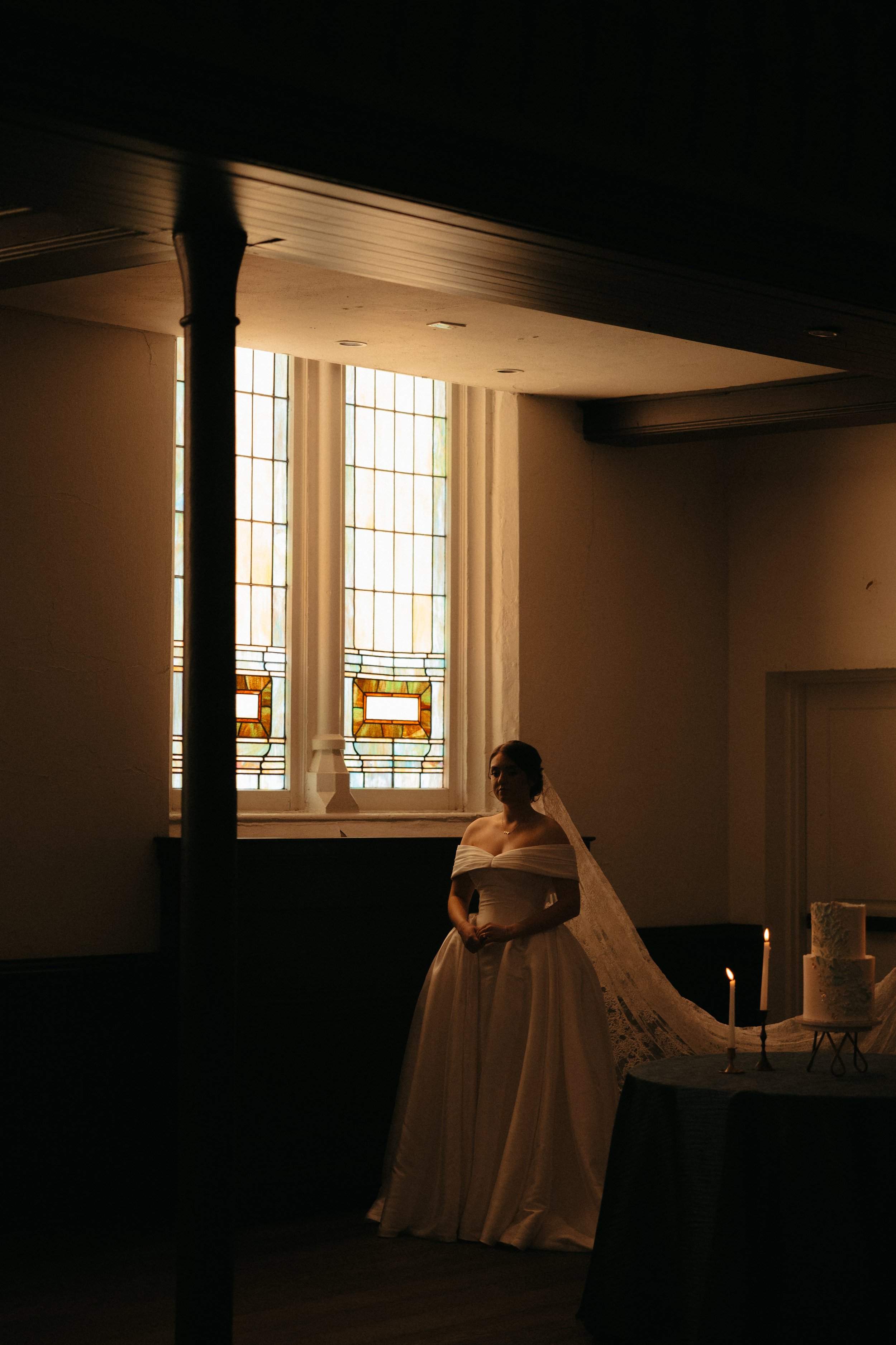 Isabel Marie Photography - A bride in a white wedding gown standing in a dimly lit room near a stained glass window with colored designs, with a wedding cake and lit candles on a table beside her.