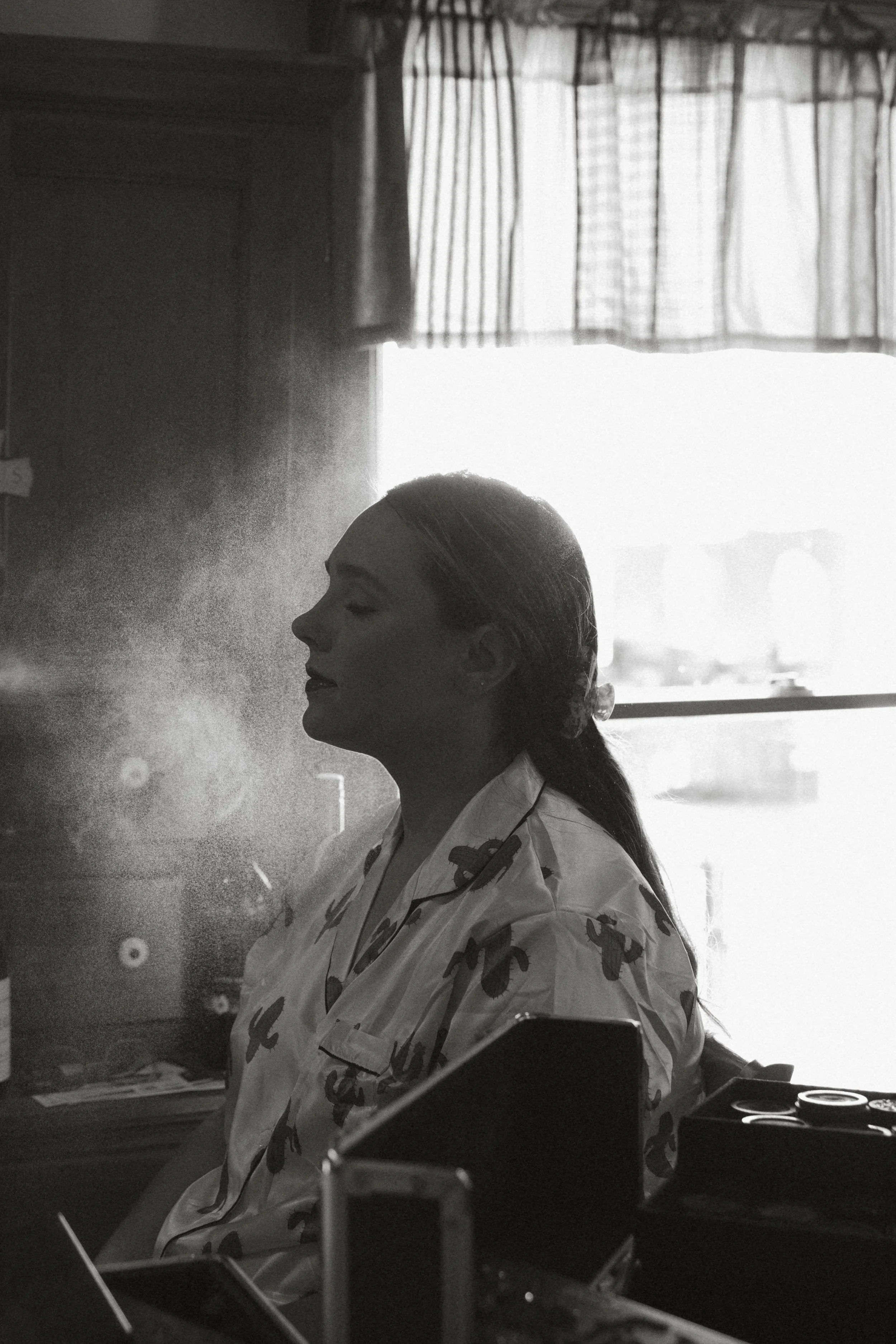 Isabel Marie Photography - A woman in pajamas sitting in a room with her eyes closed, backlit by sunlight from a window with curtains, creating a serene and peaceful atmosphere.