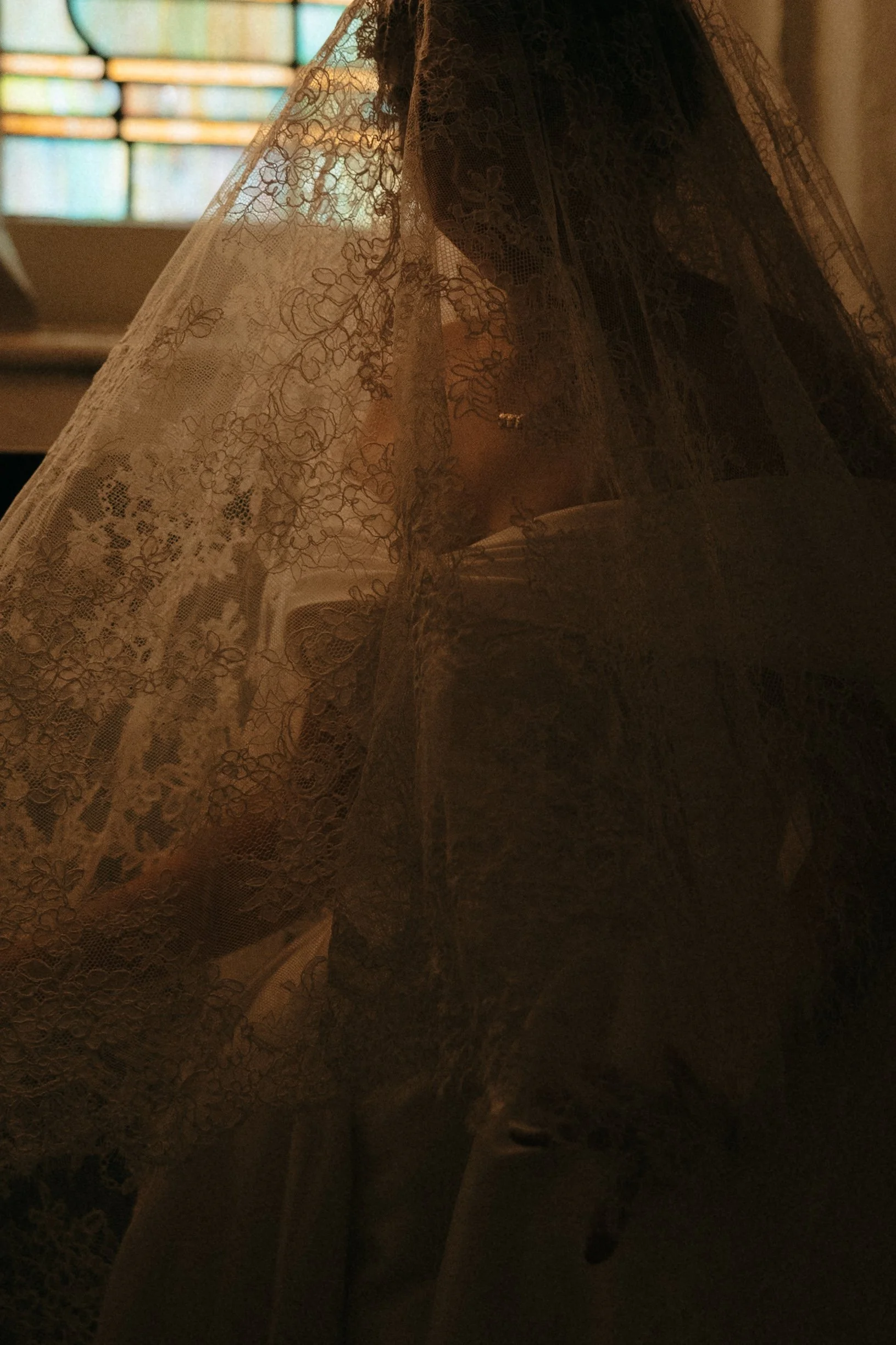 A person dressed in white clothing behind a lace veil, with only part of their face visible, in a dimly lit setting with stained glass windows in the background.