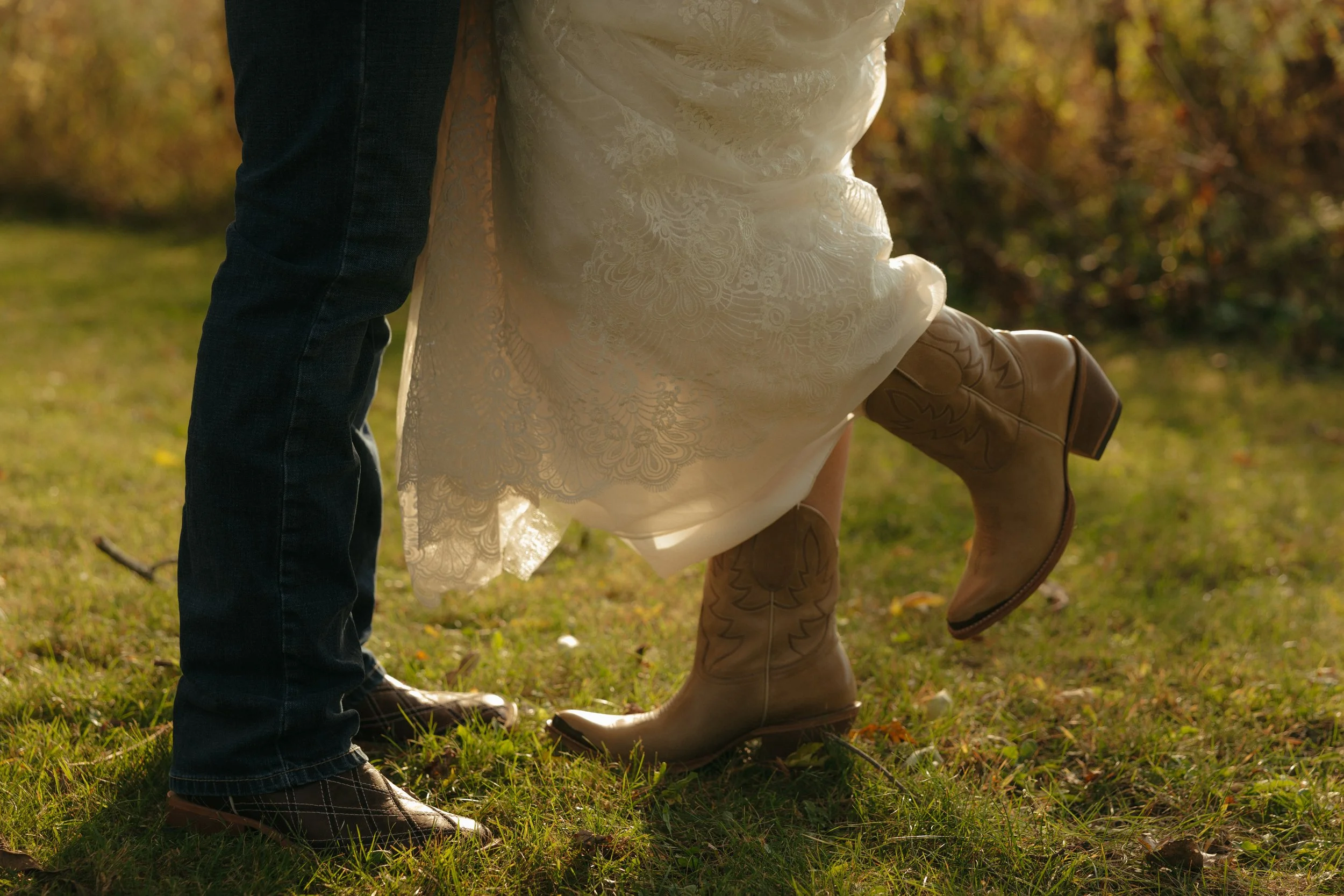 Isabel Marie Photography - A person in cowboy boots lifts a woman in a white lace dress and cowboy boots in an outdoor setting with fall foliage.