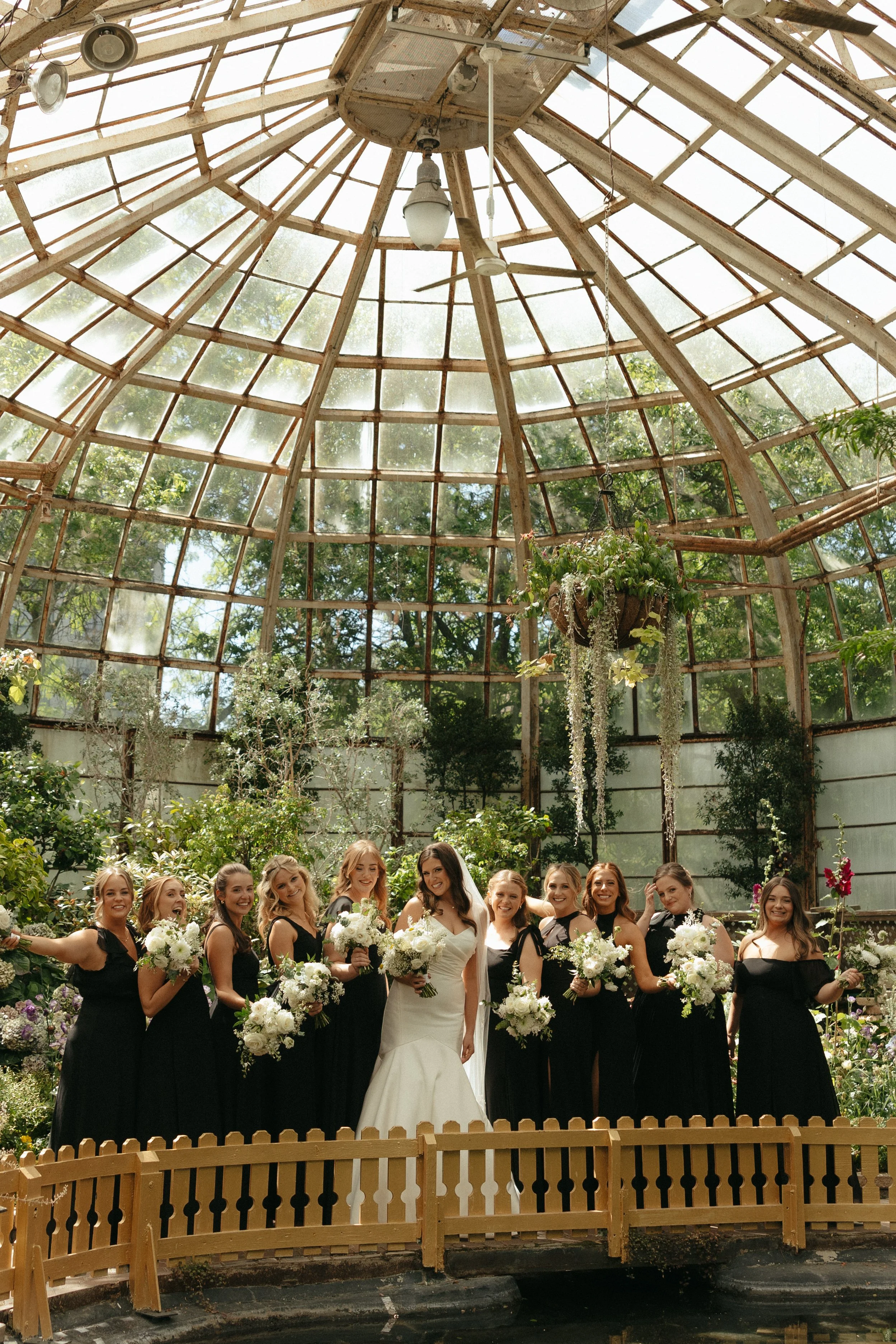 Isabel Marie Photography - A bride and ten bridesmaids standing together inside a glass greenhouse with lush greenery and hanging plants, all holding bouquets of white flowers.