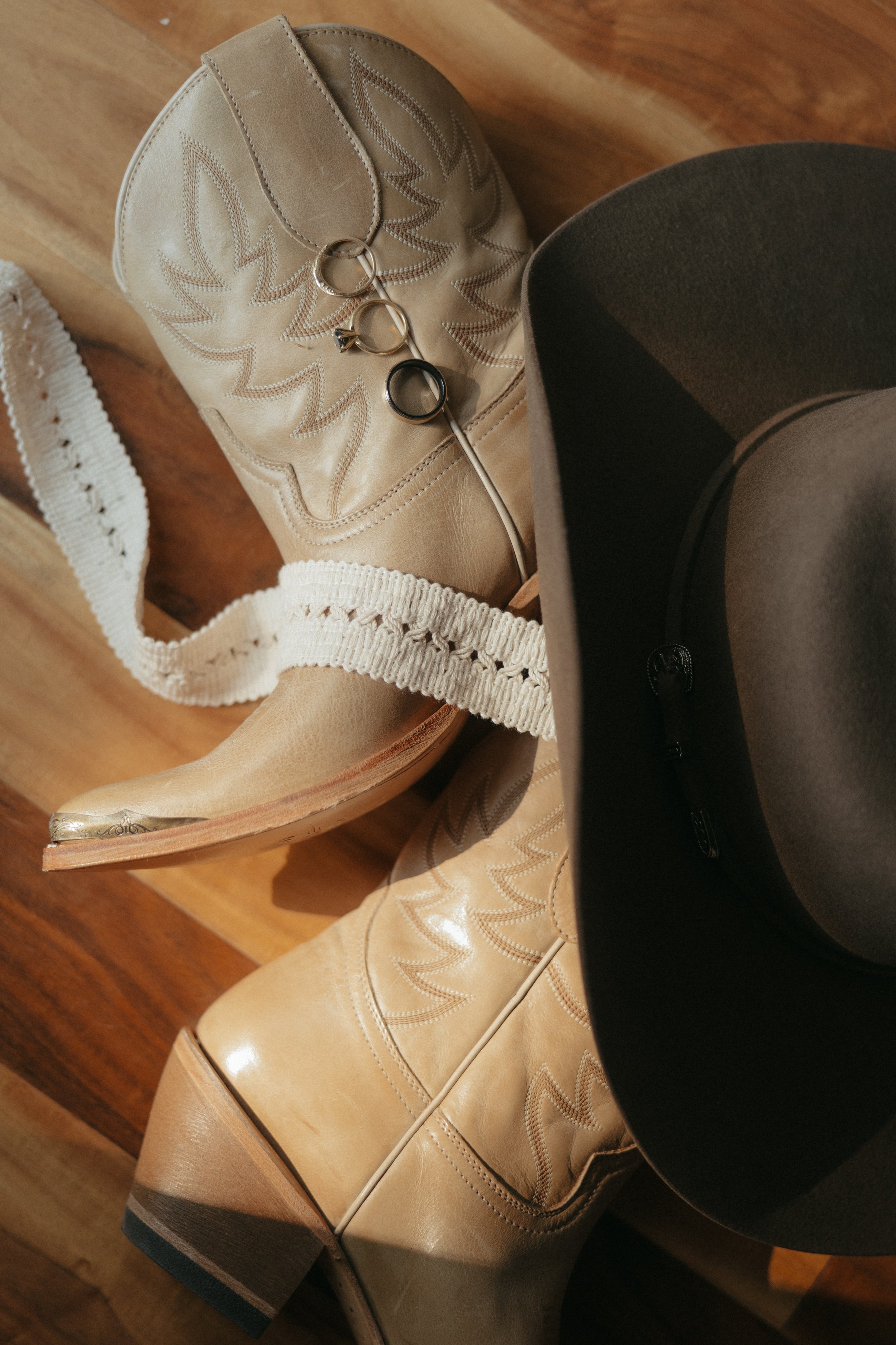Isabel Marie Photography - Flatlays A pair of tan cowboy boots, a black hat, a white lace boot strap, and multiple rings are arranged on a wooden surface.