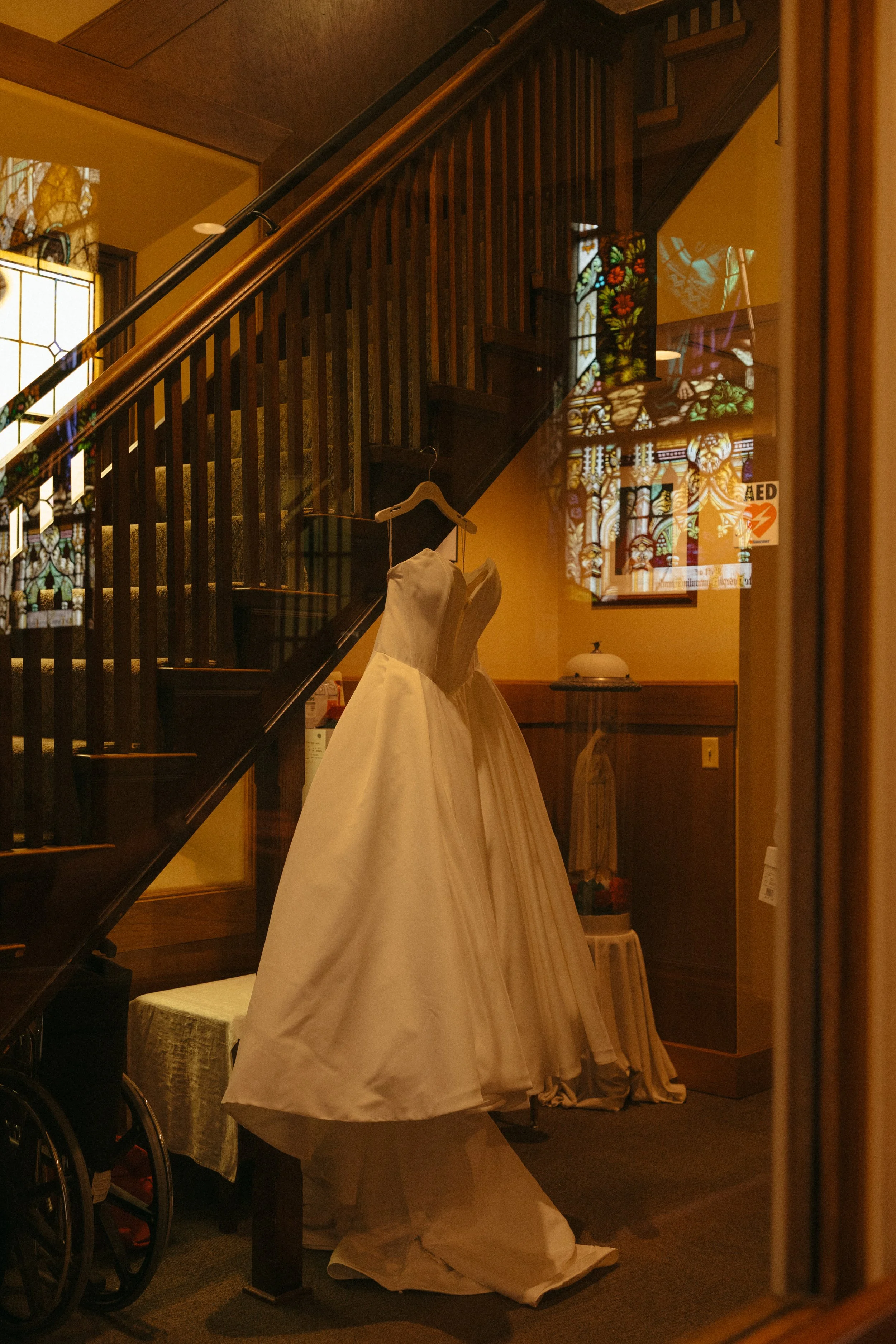 Isabel Marie Photography - A white wedding dress hanging on a wooden staircase rail inside a dimly lit room, with stained glass windows and a small table nearby.