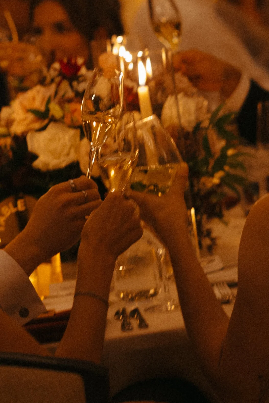 Isabel Marie Photography - People raising champagne glasses in a toast at a celebration with a floral arrangement and lit candles in the background.