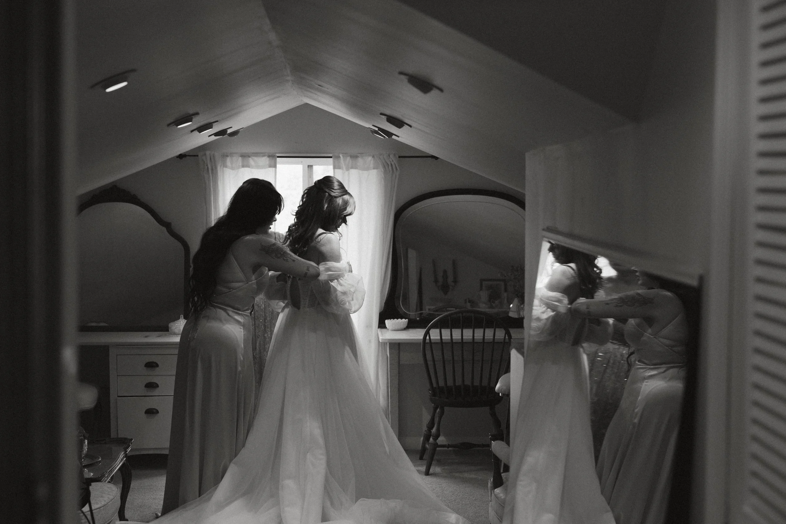 Isabel Marie Photography - Two women, one in a wedding gown and the other helping her, in a bedroom with a window and mirror, in black and white.