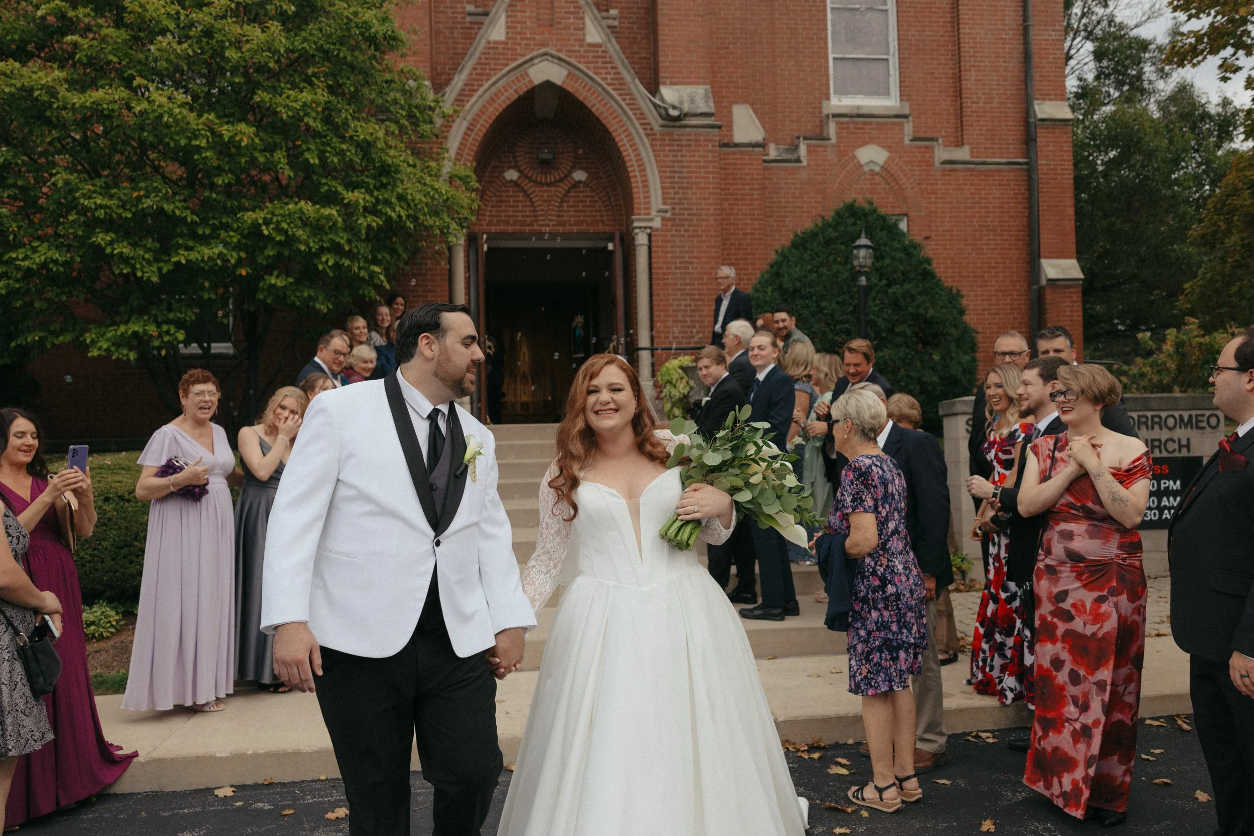 Isabel Marie Photography - A newlywed couple is smiling and holding hands outside a brick church, surrounded by friends and family celebrating their wedding.