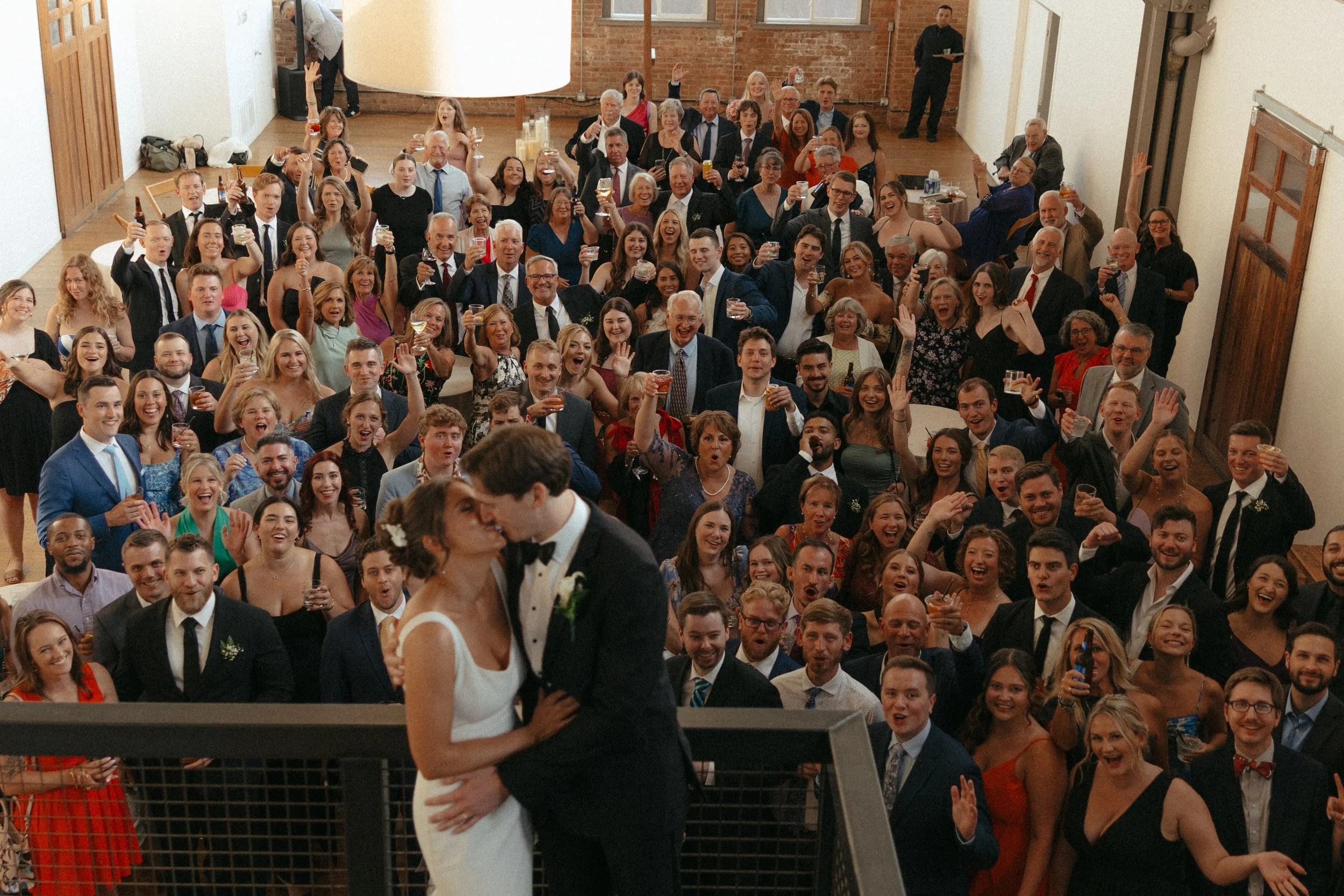 Isabel Marie Photography - A wedding celebration showing a bride and groom kissing in front of a large crowd of guests holding drinks and celebrating in a reception hall.