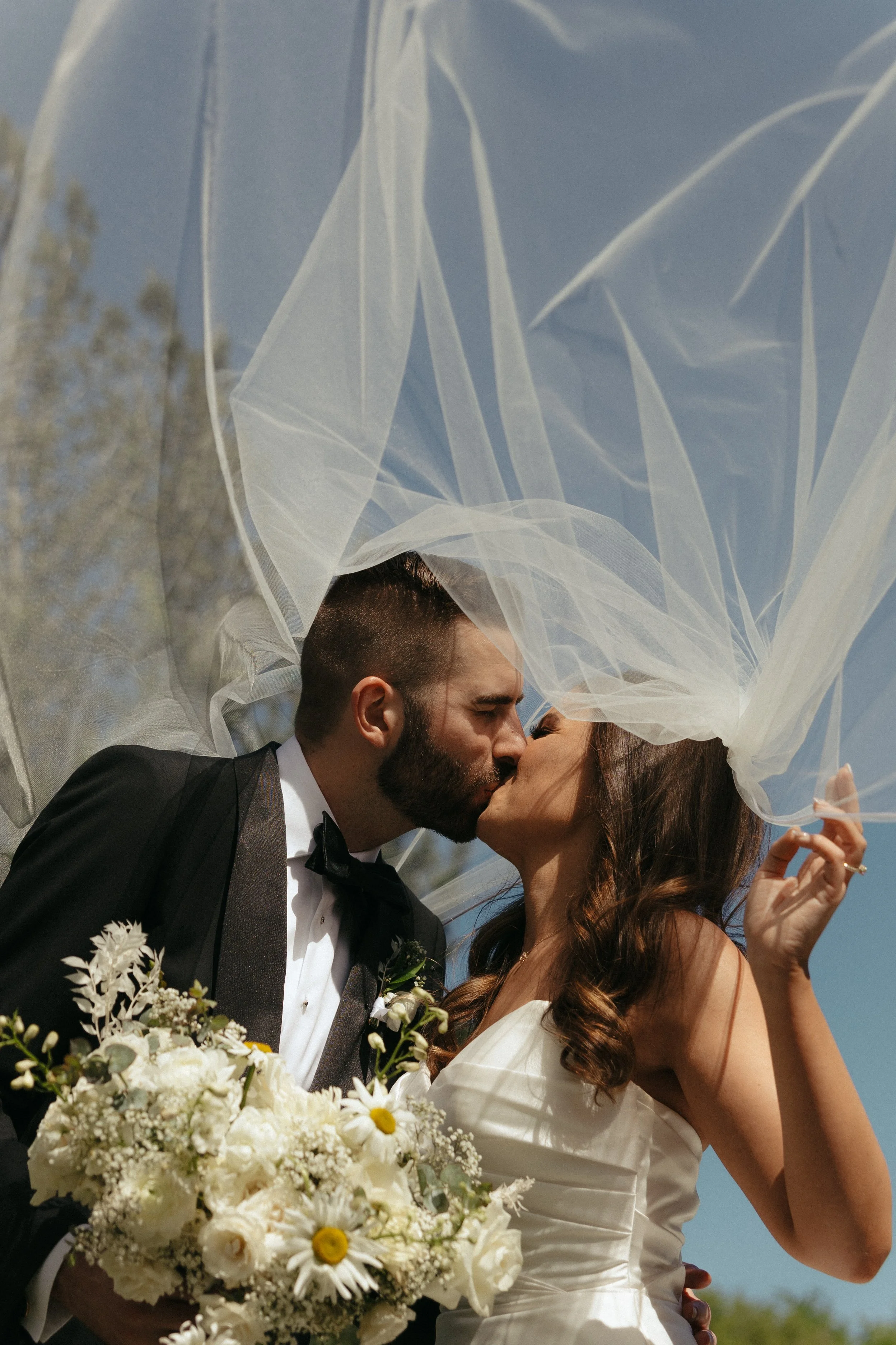 Isabel Marie Photography - A bride and groom kissing under a sheer veil during their outdoor wedding, with the bride holding a bouquet of white flowers.