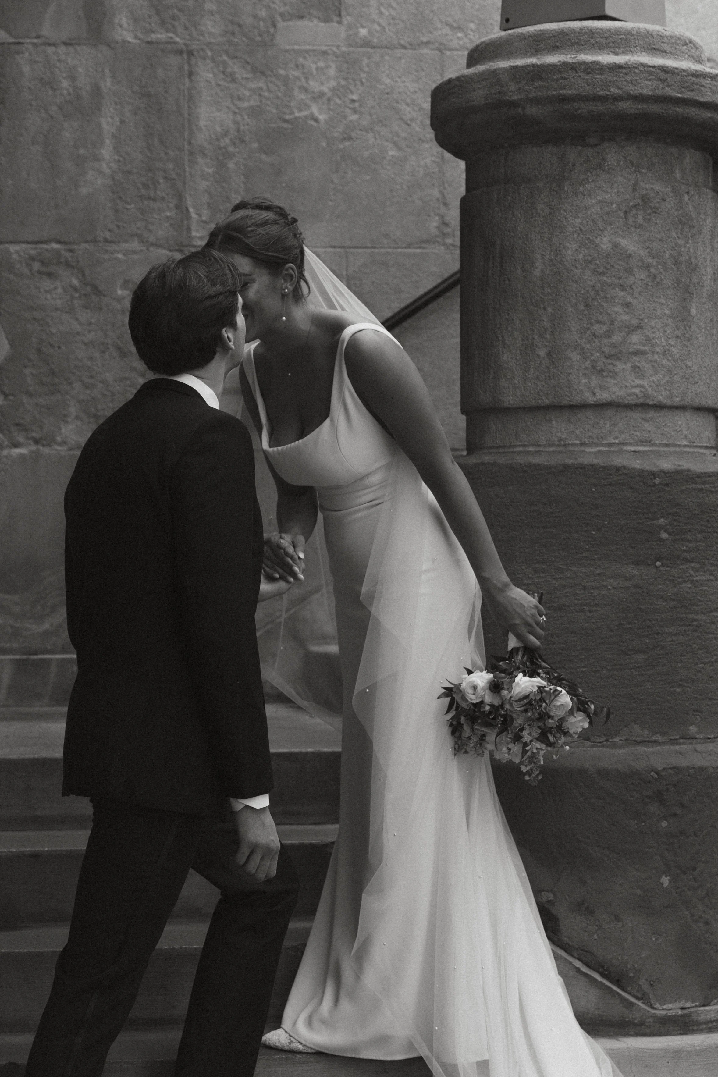 Isabel Marie Photography - A black-and-white photo of a newlywed couple sharing a kiss, with the bride holding a bouquet, standing on stairs next to a stone wall and a large stone pillar.