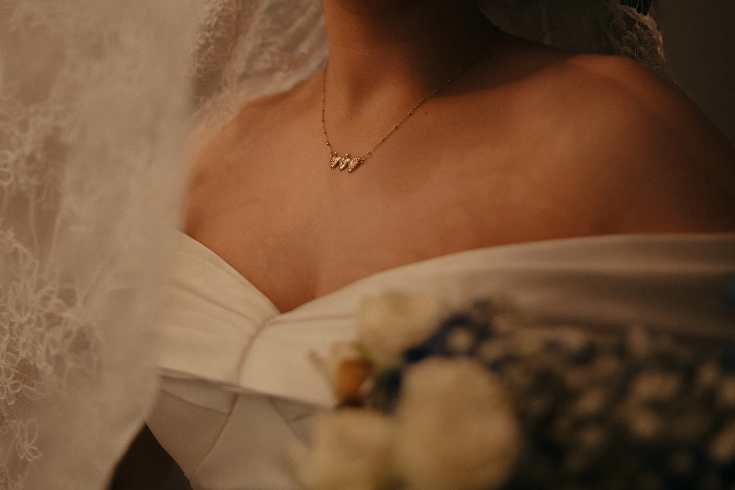 Isabel Marie Photography - Close-up of a woman's shoulder and décolletage, showing a gold necklace with multiple small triangular pendants, dressed in a white off-shoulder gown with lace detailing on the sleeve.
