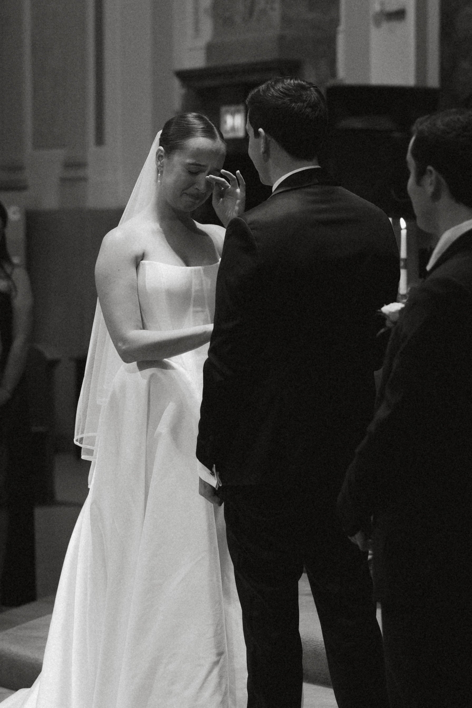 Isabel Marie Photography - A black and white photo of a bride and groom during their wedding ceremony inside a church. The bride is crying and touching her nose, while the groom and another man stand nearby.
