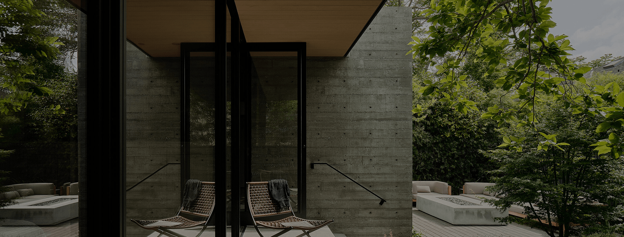 Modern house exterior with concrete walls, black metal and glass doors, outdoor seating, surrounded by trees and greenery.