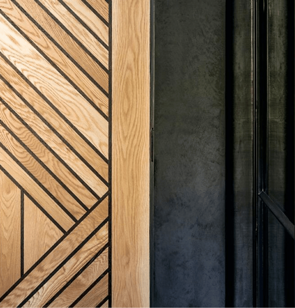 Close-up of wooden panels with geometric pattern and black lines on the left, and a dark wall with a metal frame or panel on the right.