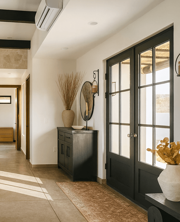 Interior view of a house entrance with black double doors, a sideboard with decorative items, large vase with dried plants, a mirror, and a light fixture, with sunlight coming through the glass doors.