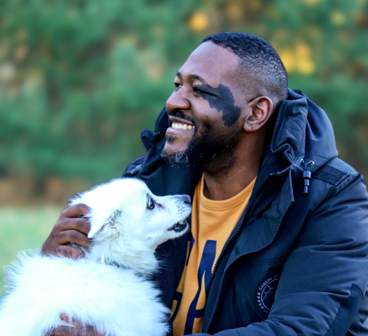 A man with a beard and short hair, wearing a black jacket and yellow shirt, smiling and holding a white puppy with blue eyes near his face in an outdoor park setting.