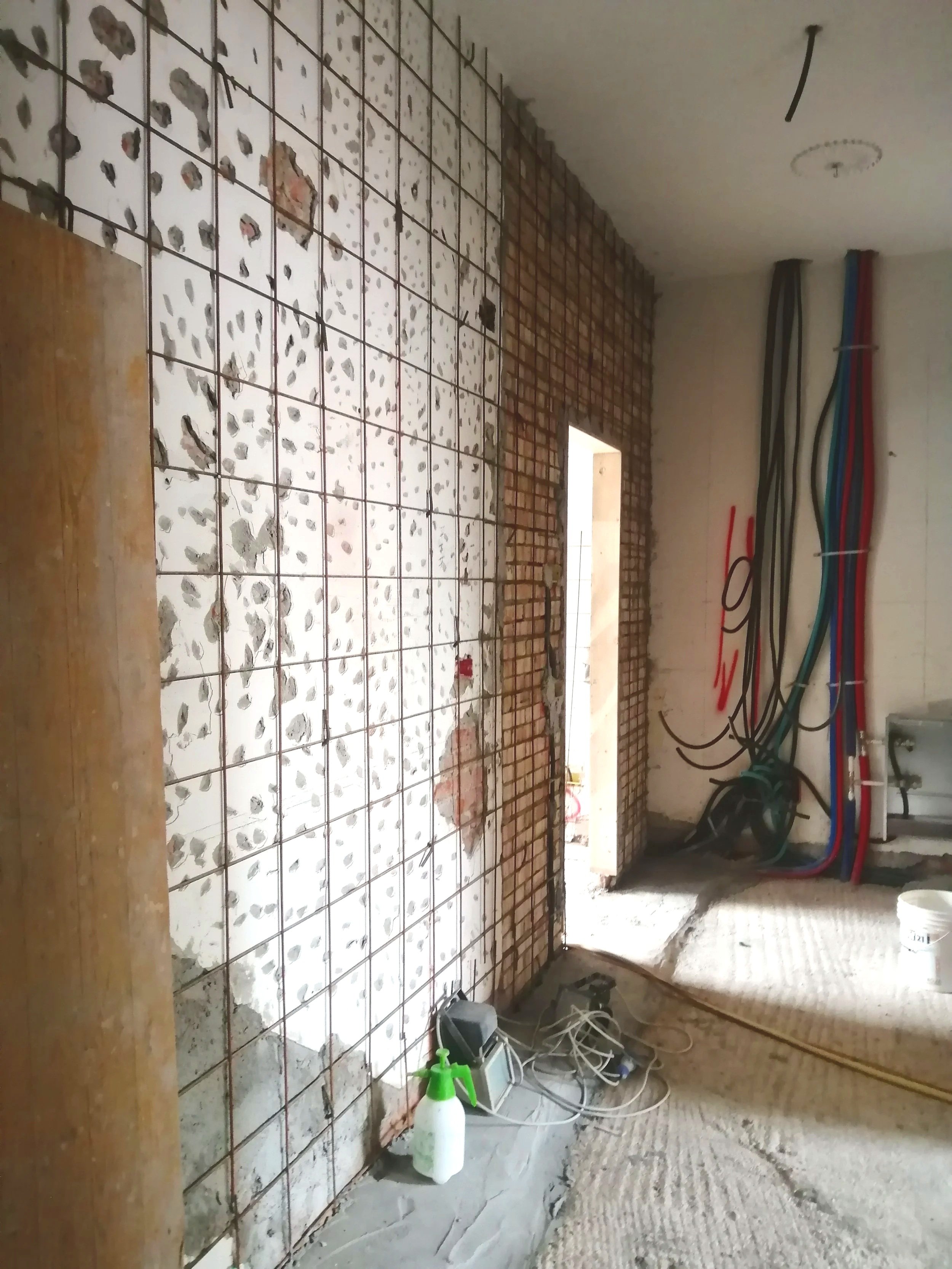Interno di una stanza in fase di ristrutturazione con pavimento irregolare, muro con reti metalliche e mattonelle, e cavi elettrici appesi alla parete. Vari attrezzi e materiali per lavori edili sono presenti sul pavimento.