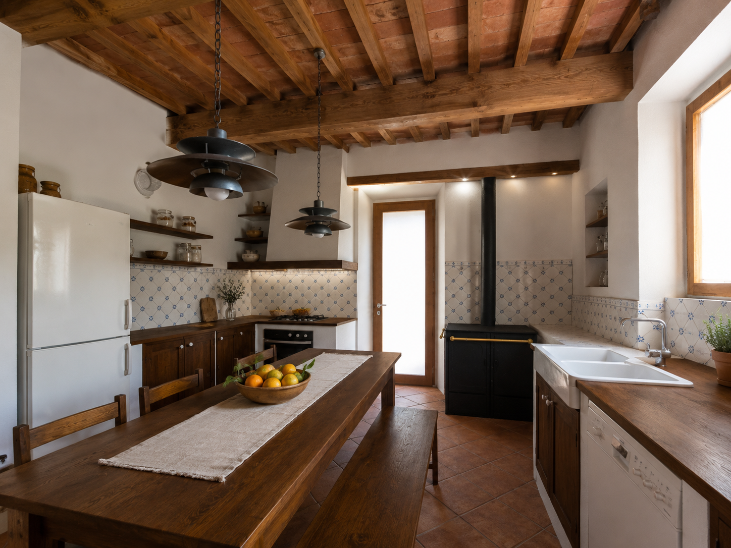Cucina rustica con soffitto in legno, tavolo di legno con sedie, frutta in una ciotola, cucina con forno, lavello, stoviglie in vetro su scaffali, camino nero, pareti con piastrelle bianche con motivo blu, finestra grande, porta di vetro. 
