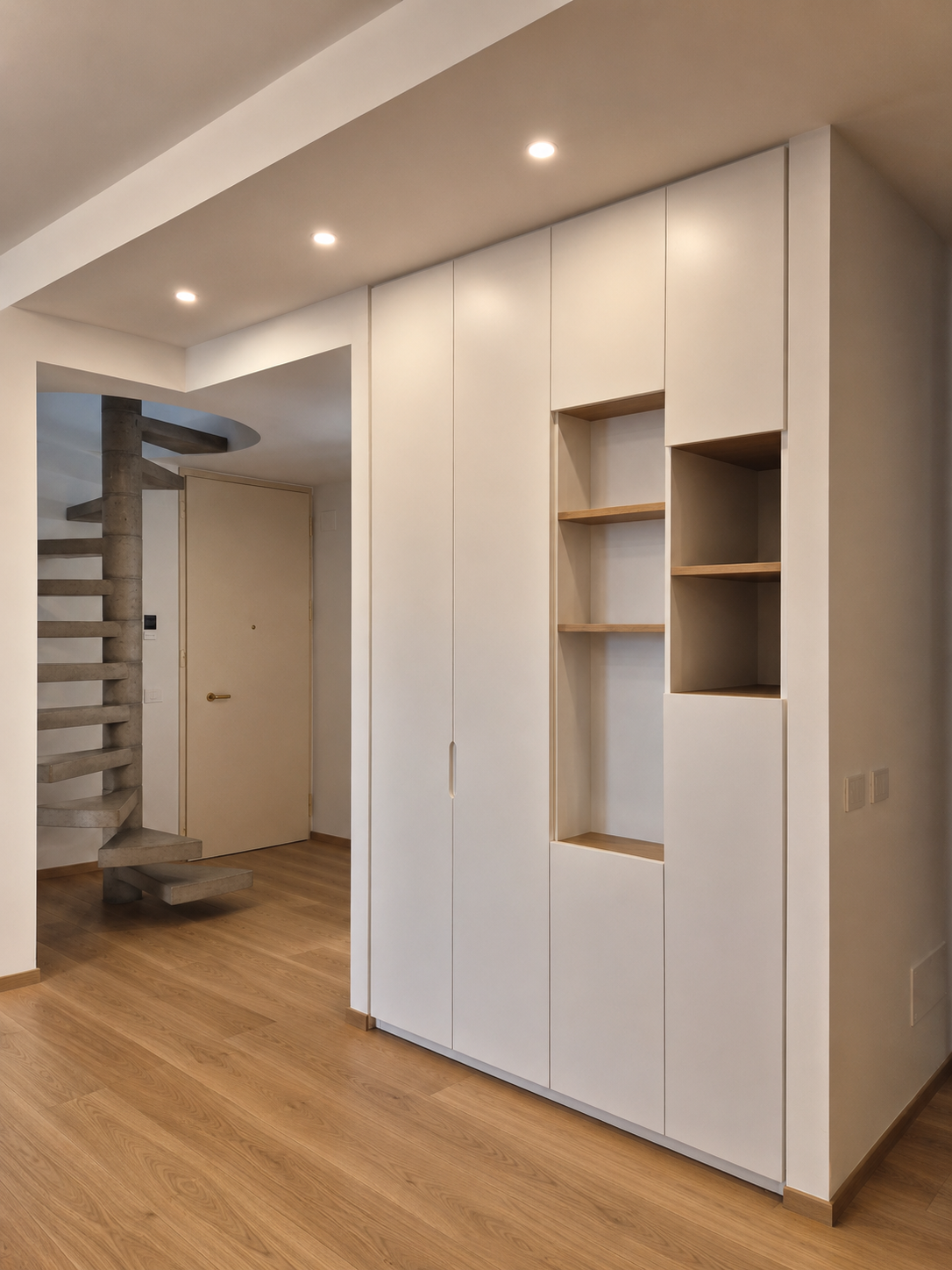 Interno di un appartamento con pareti bianche, pavimento in legno chiaro, e armadio bianco con mensole di legno. In fondo si vede una scala a spirale in cemento grigio e una porta beige.