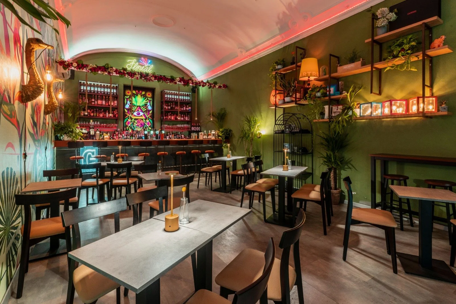 Interno di un bar o ristorante con pareti verdi, arredamento moderno, tavoli con candele, scaffali con piante e decorazioni, bar sullo sfondo con neon e murale colorato