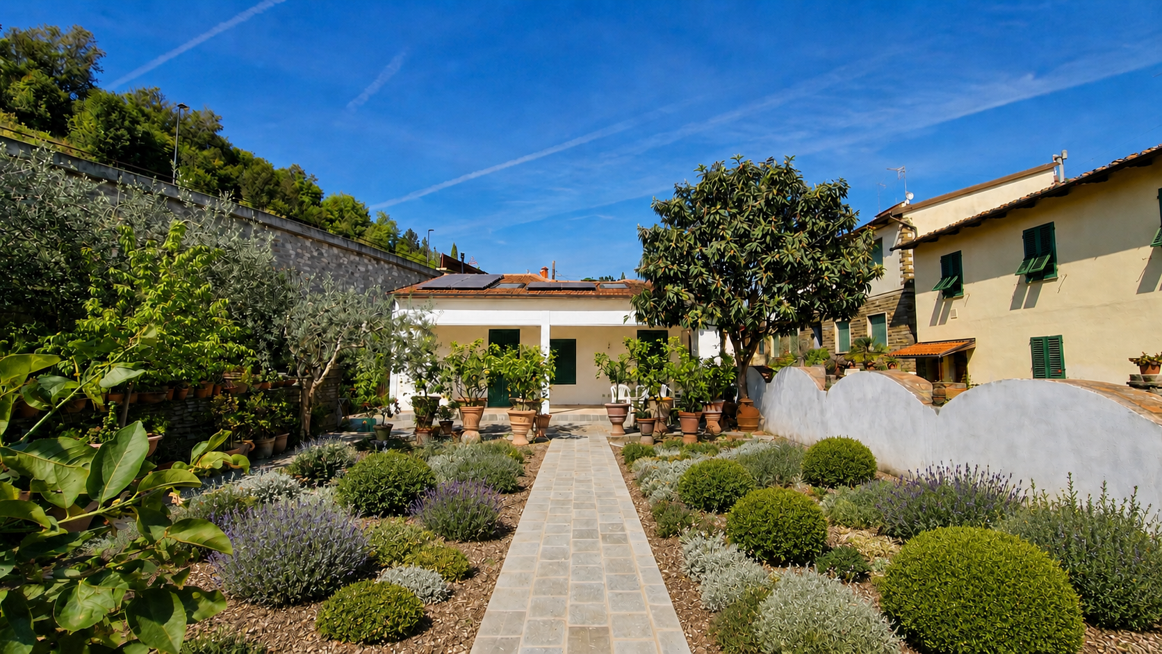 Giardino con piante e arbusti fioriti, sentiero centrale, case con finestre verdi e pareti bianca e gialla, cielo azzurro con nuvole