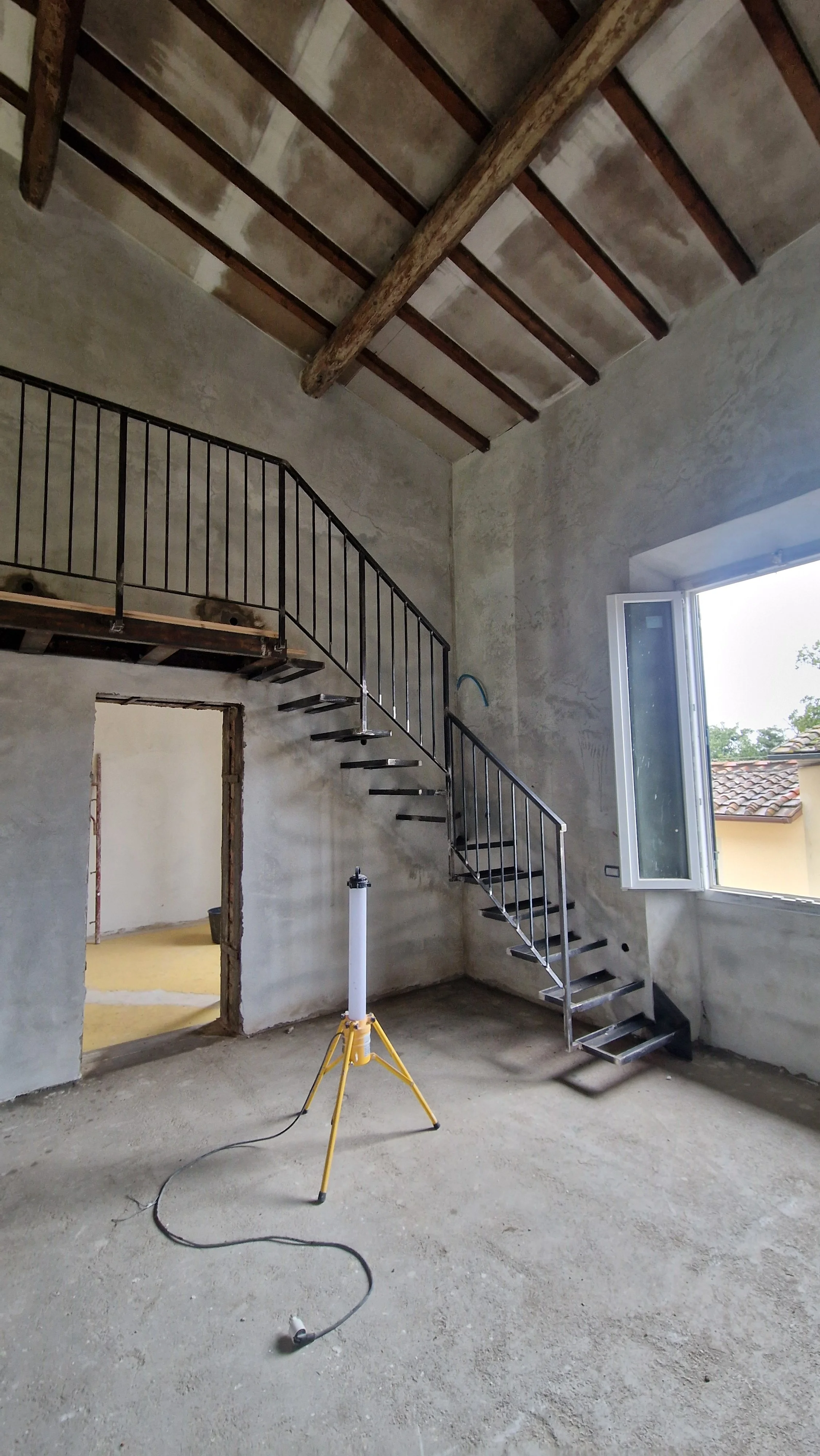 Interno di una casa in costruzione con scala metallica, pareti di cemento non verniciate, un grande finestrone aperto, e un lampada di sicurezza sul pavimento