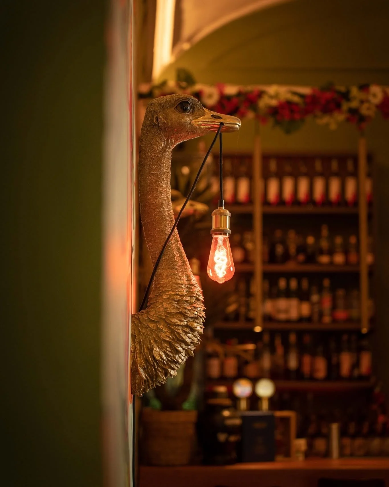 Lampada decorativa appesa al muro con lampadina a incandescenza accesa appesa al becco. Sfondo di scaffali con bottiglie e oggetti. Atmosfera calda e intima.