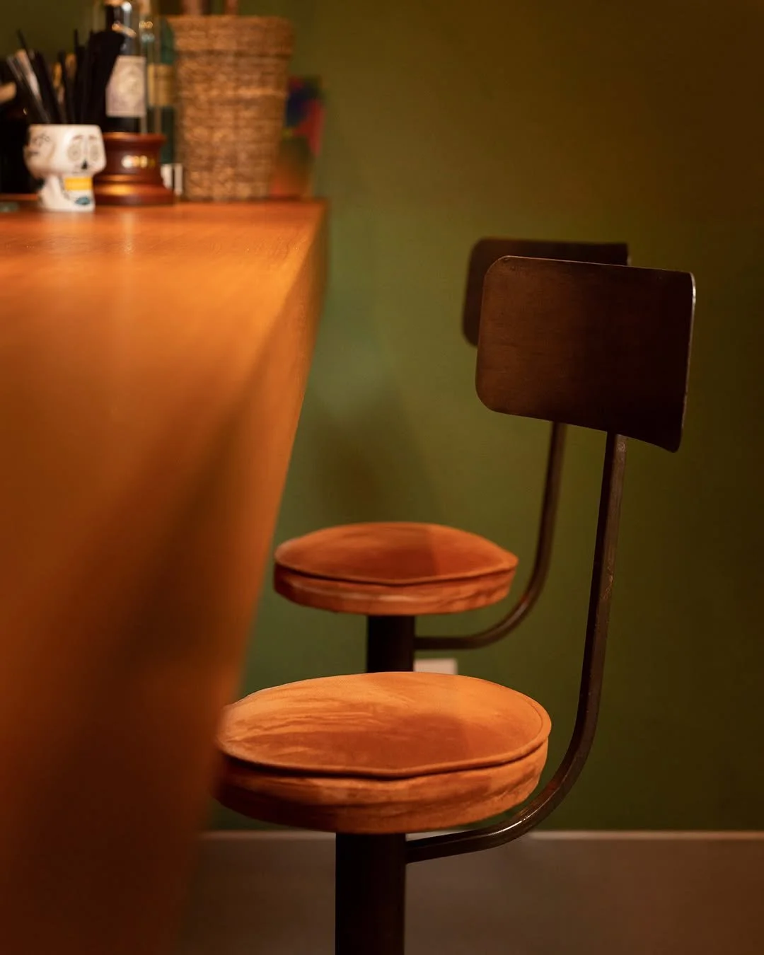 Angolo di un bar con due sedie di legno e un bancone di legno. Sul bancone ci sono oggetti vari, tra cui un cestino di vimini e contenitori.