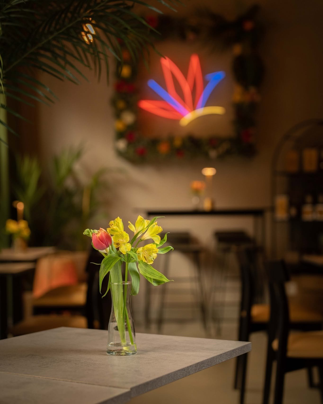 Un vaso con fiori colorati, tra cui tulipani e alstroemerie, su un tavolo in un ambiente di ristorante o caffetteria decorato con luci colorate e una corona natalizia sul muro in background.