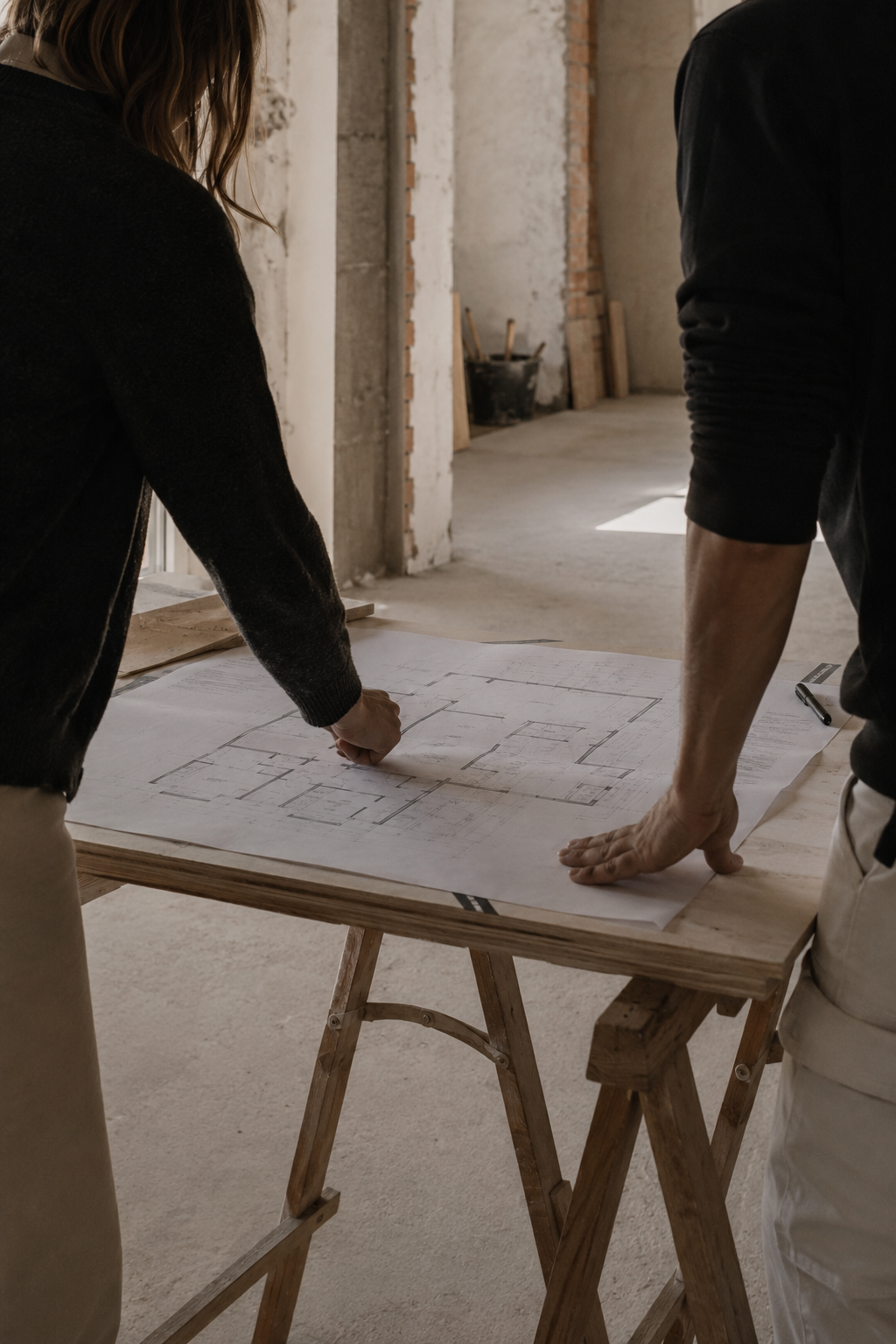 Due persone stanno esaminando un progetto architettonico su un tavolo di legno in un cantiere o sede di un cantiere, con pareti in mattoni e cemento.