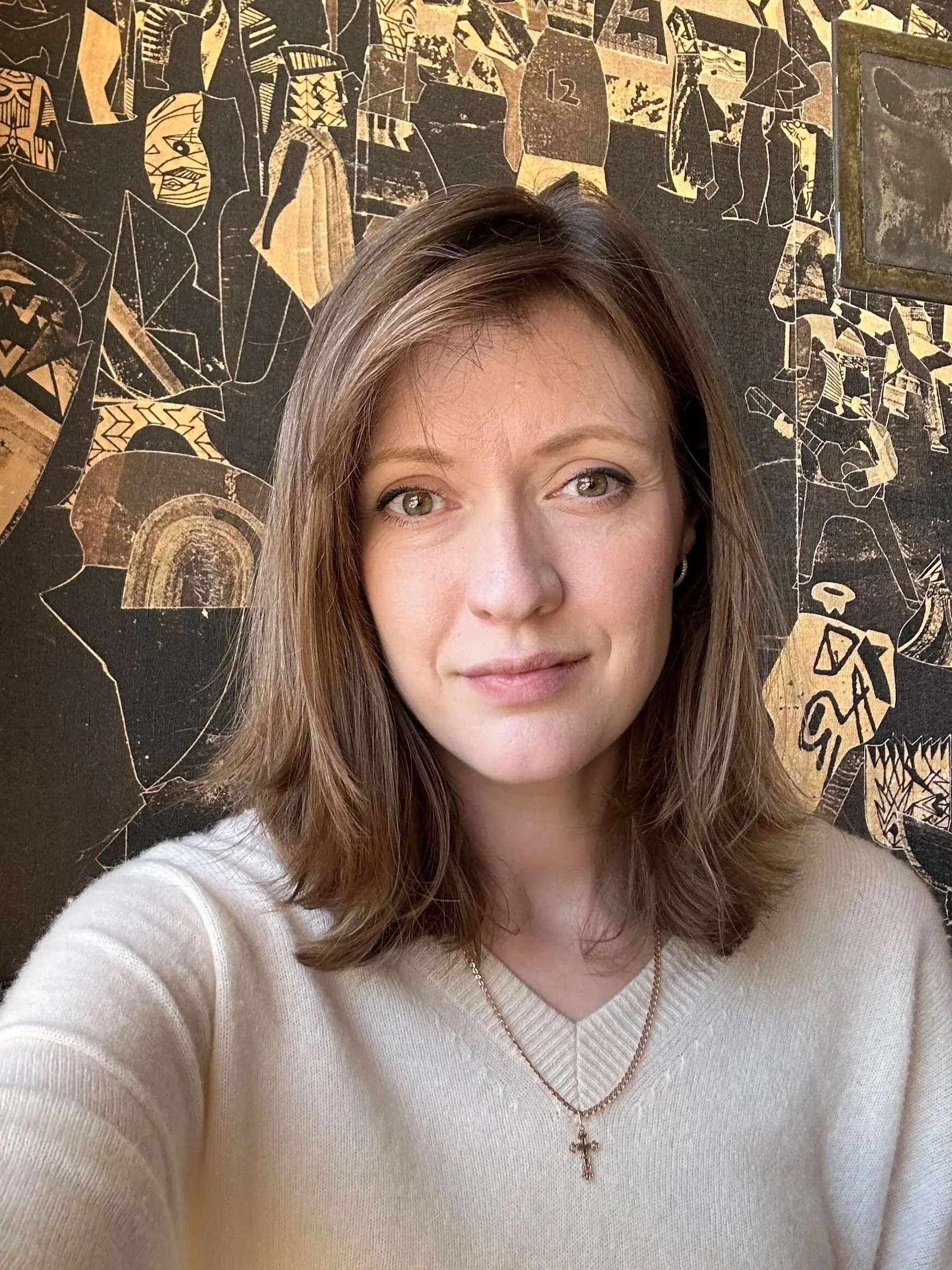 A woman with light brown hair and hazel eyes taking a selfie in front of a decorative wall with black and gold abstract art, wearing a beige sweater and a necklace with a cross pendant.