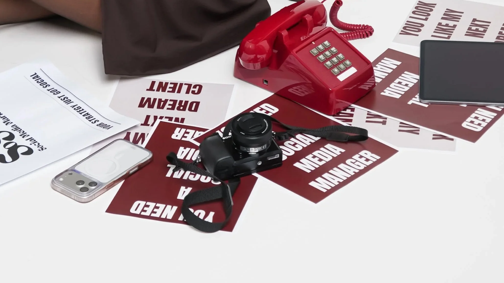 A cluttered white table with various items including a red rotary phone, a black digital camera, a smartphone in a clear case, a sheet of paper, a brown bag, and several signs with warning and media text.
