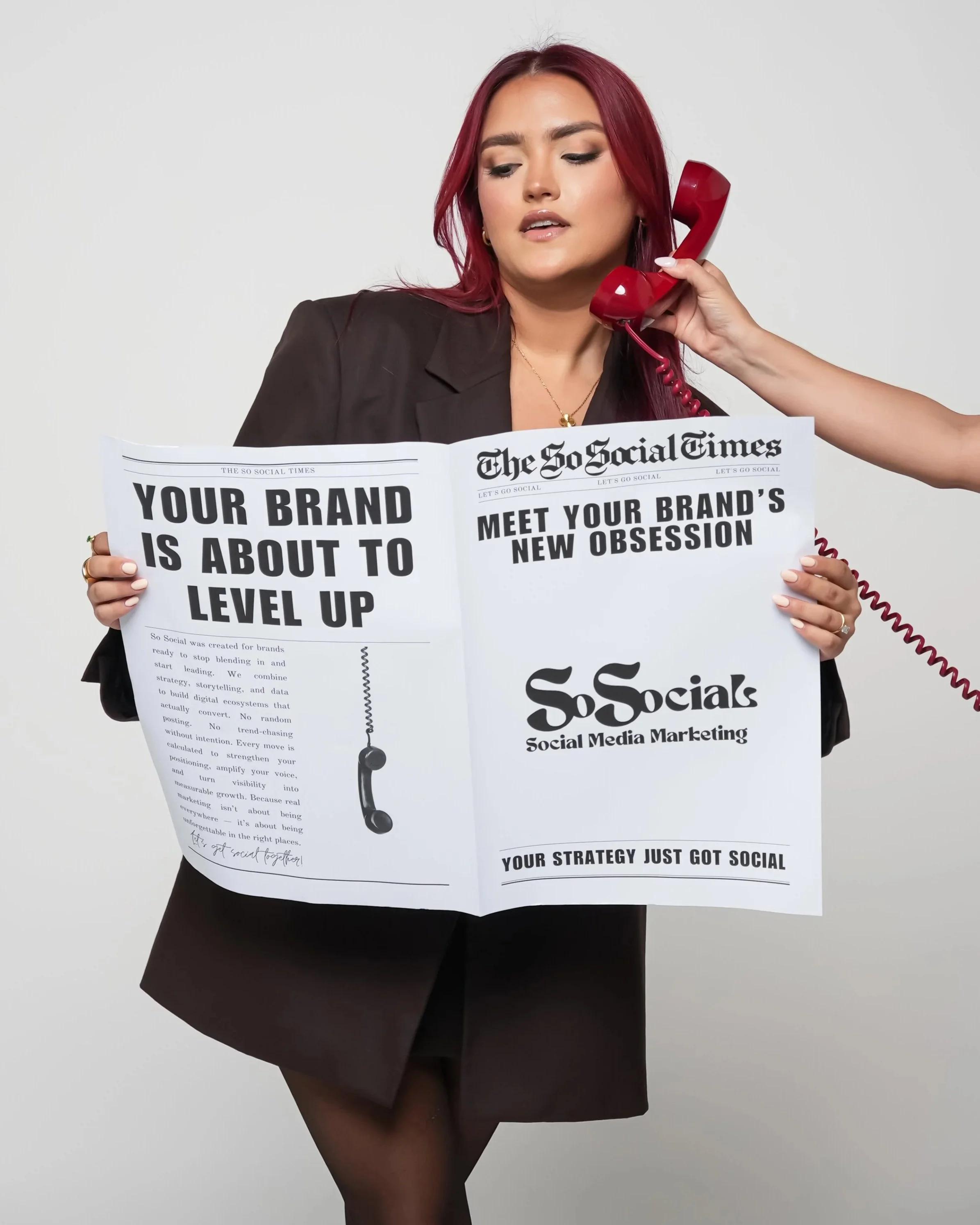A woman with red hair in a black blazer holding a large flyer about social media marketing, while talking on a red rotary phone.