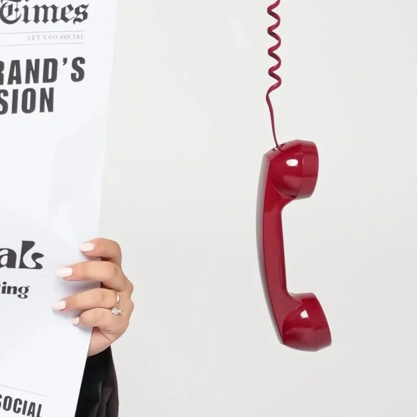 A red vintage rotary phone hanging from the ceiling with a pink curly cord, partially obscured by a person holding a newspaper.