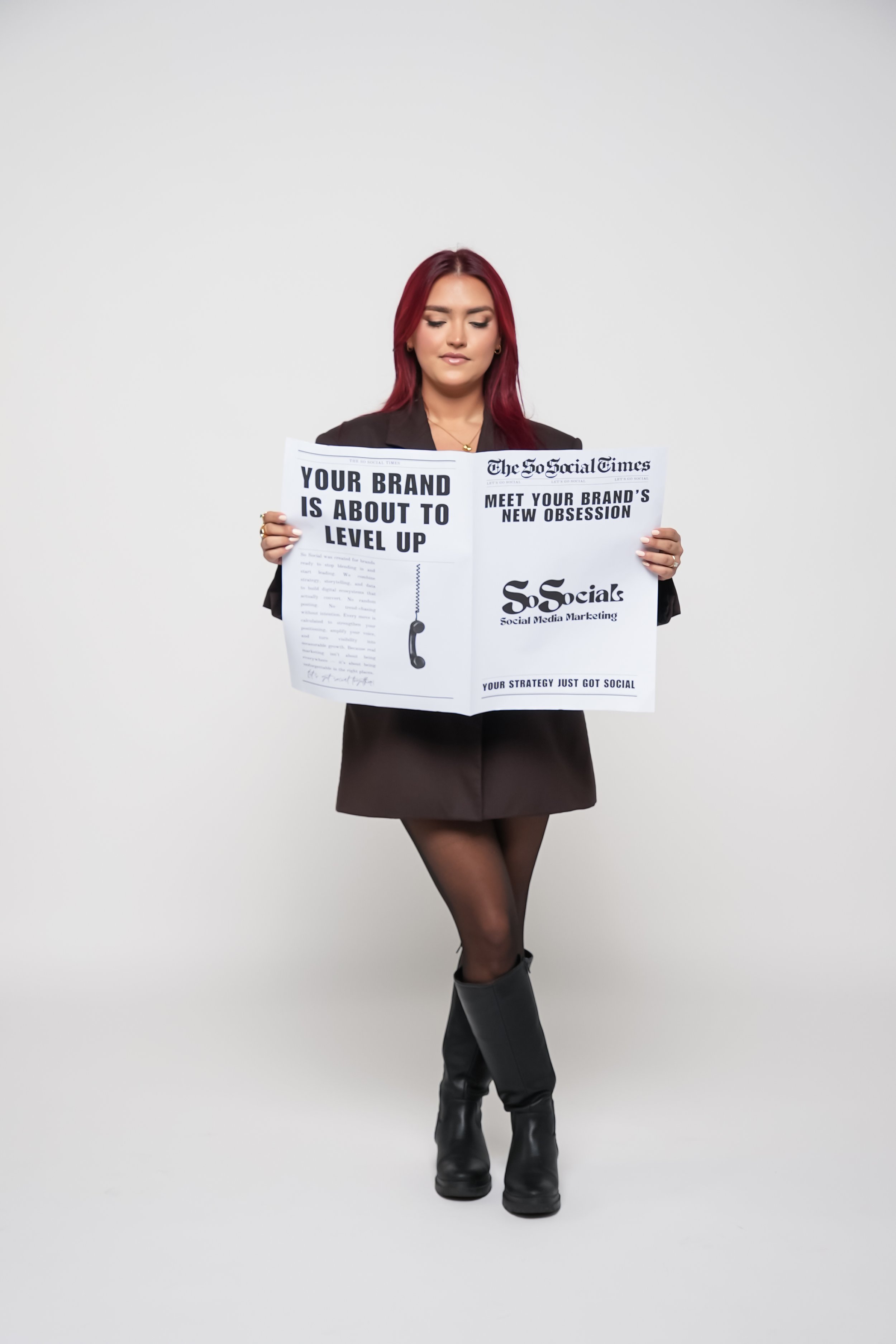A woman with long red hair wearing a black blazer, black skirt, stockings, and knee-high boots is holding a newspaper that promotes social media marketing strategies.