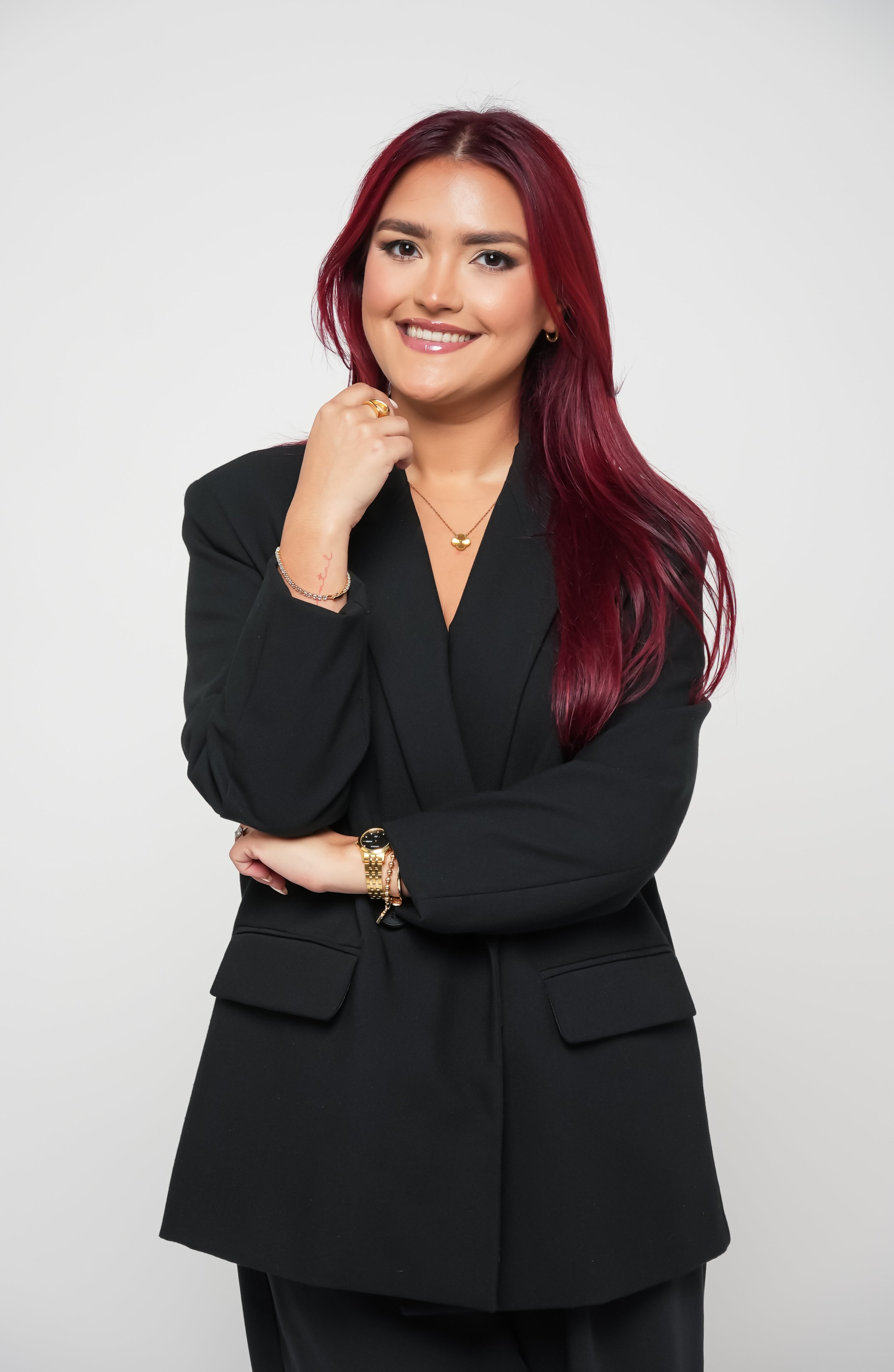 A woman with long red hair, dressed in a black blazer, smiling and posing with her right hand near her face, against a plain white background.