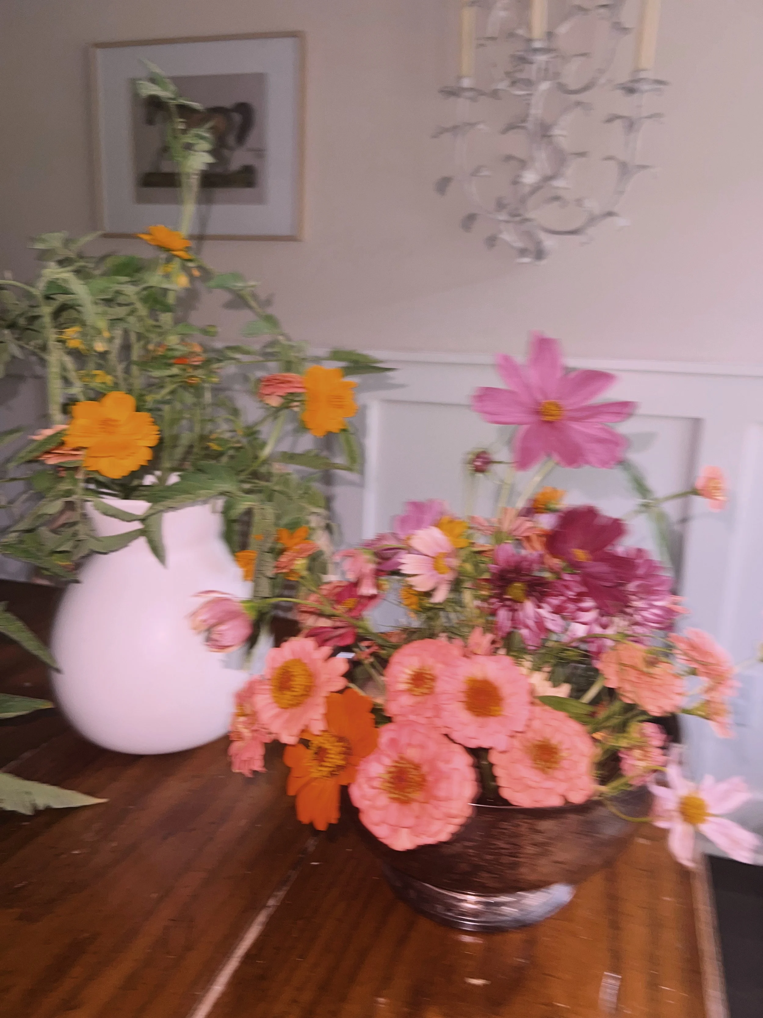 Vase filled with pink and orange flowers on a wooden surface, with framed art and a chandelier on the wall in the background.