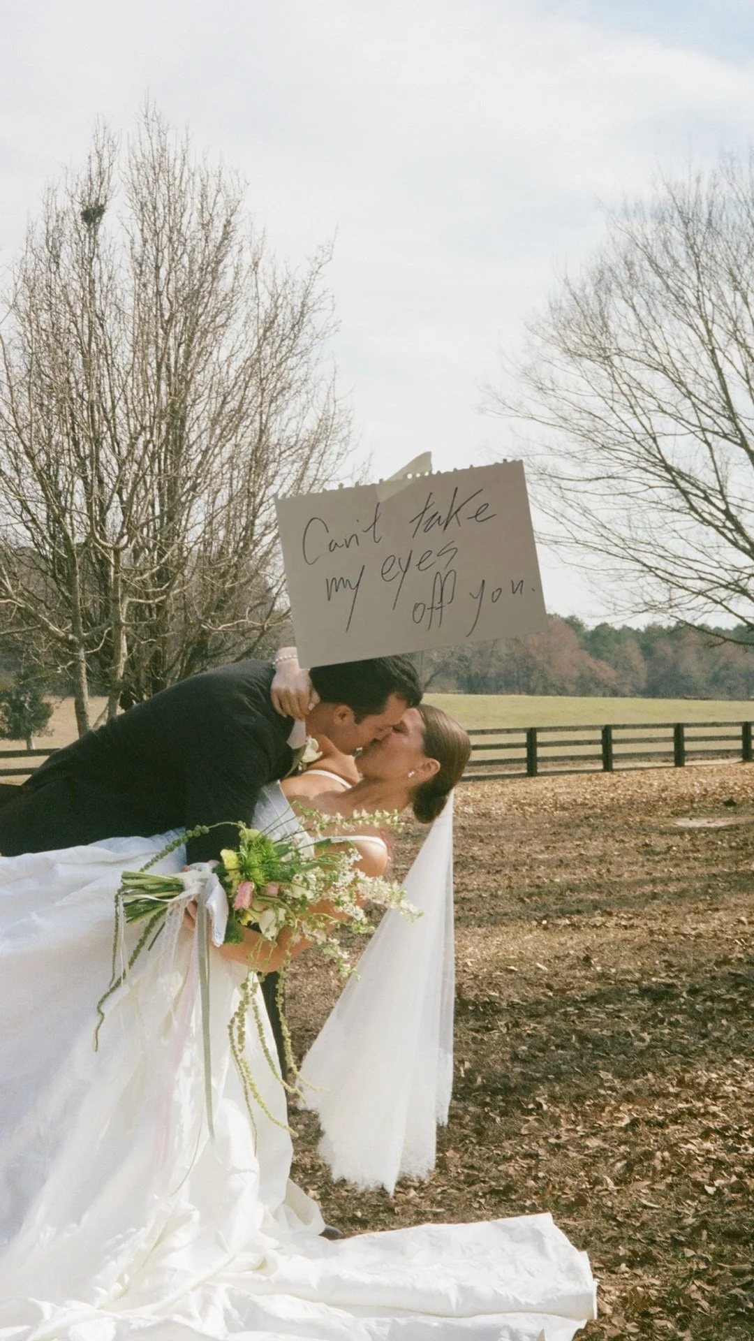 A bride and groom kiss outdoors on their wedding day, holding a bouquet of flowers. A man above them holds a sign that reads "Can't take my eyes off you." Bare trees and a fence are in the background.