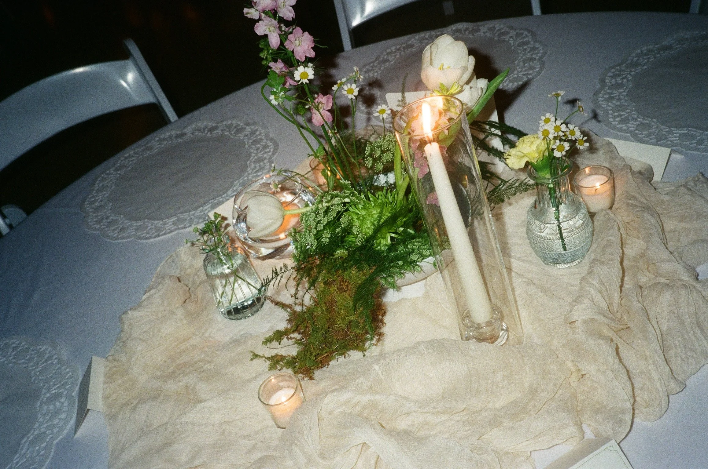 A table decorated with flowers, candles, and lace, featuring a centerpiece with pink and white flowers in a glass vase and gold candlesticks.