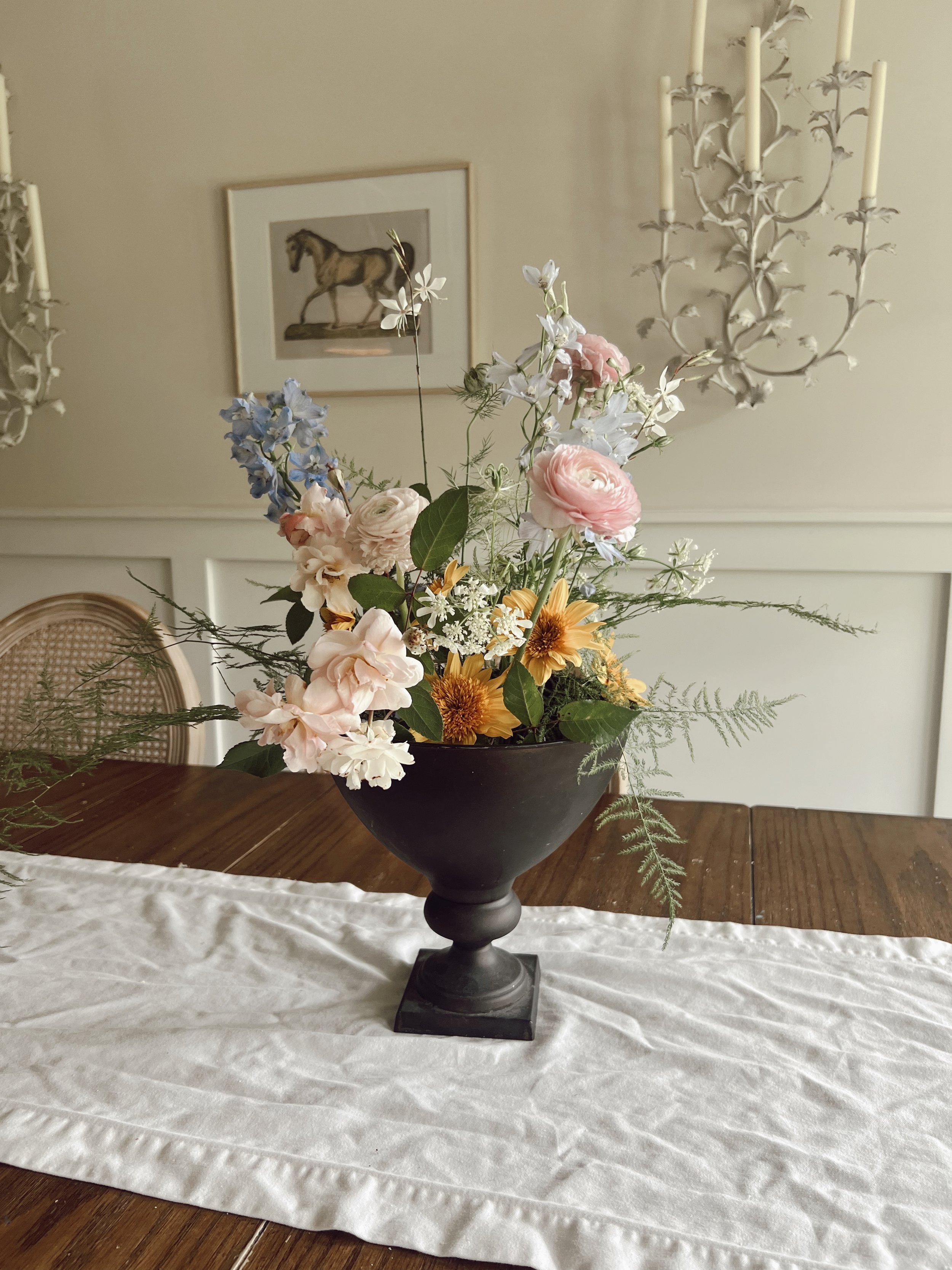 A black vase with a colorful flower arrangement including roses, sunflowers, and various other flowers and greenery, placed on a white tablecloth on a wooden table. In the background, there is a wall with a framed picture of a horse and an ornate cha