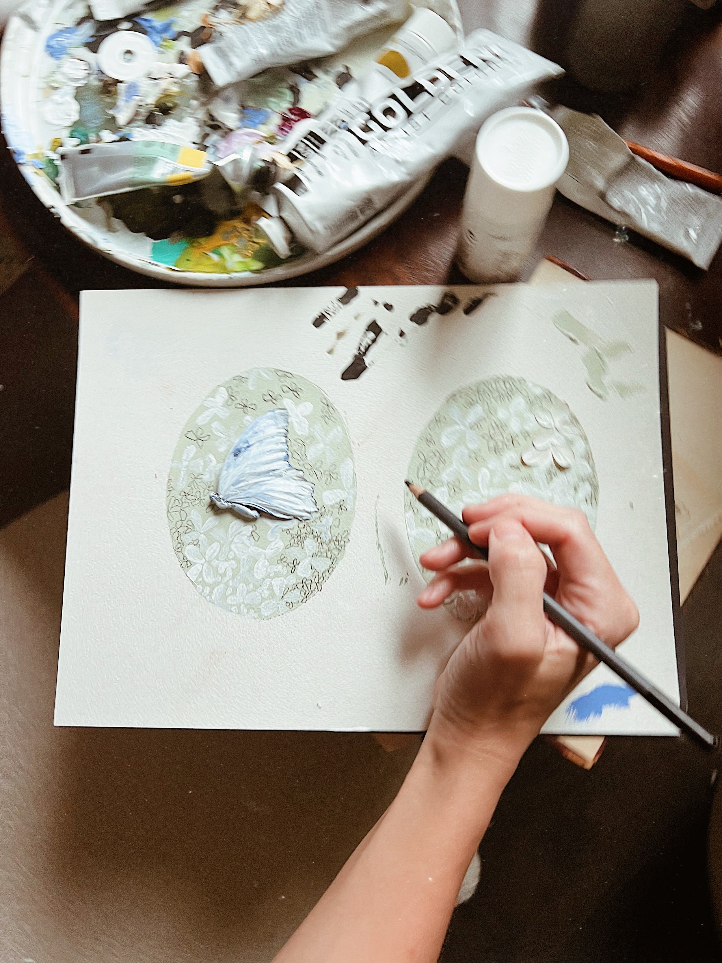 Person painting a butterfly on a canvas, with a palette of colors nearby and art supplies on the table.