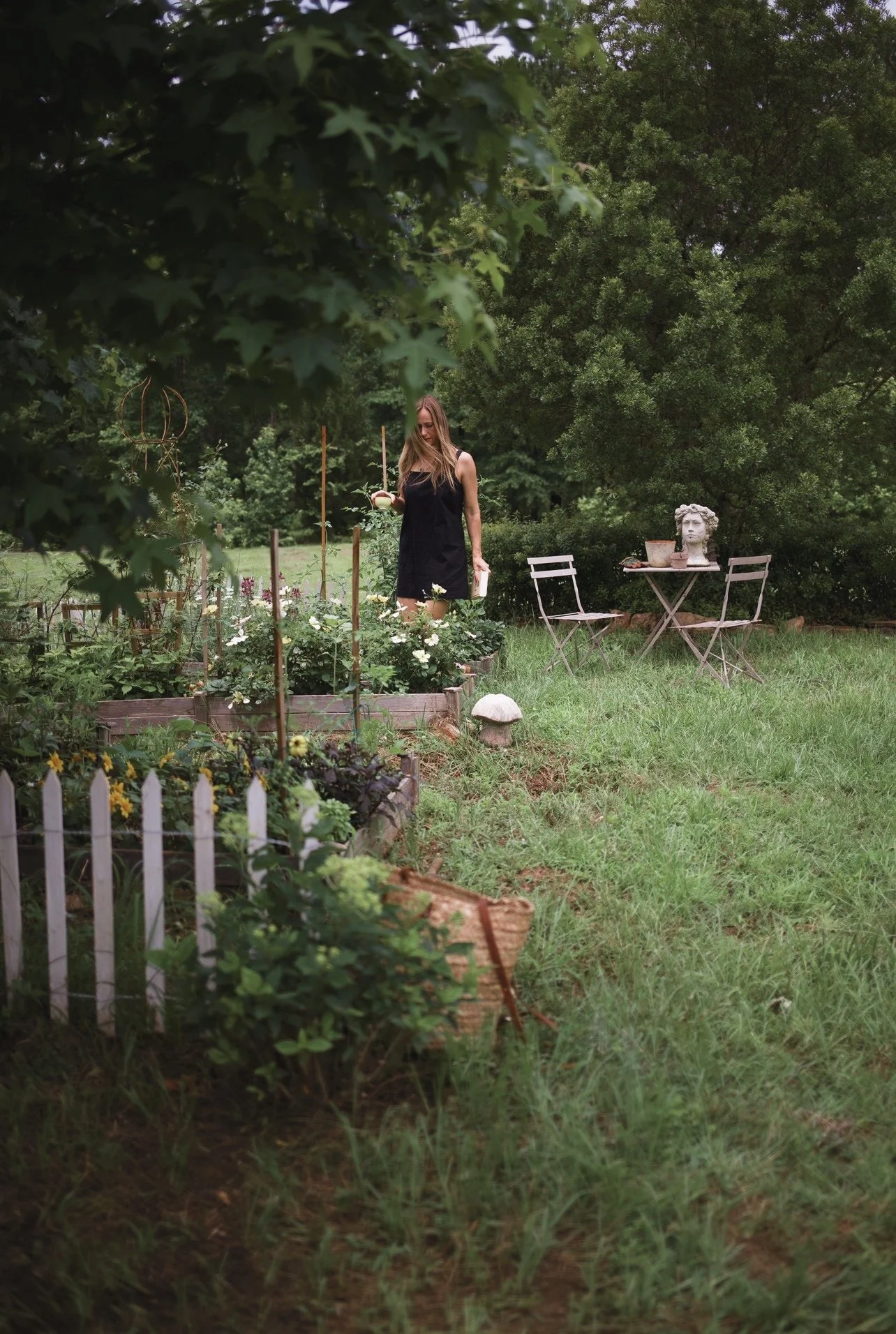 A woman in a black dress watering plants in a garden with lush green trees and a small patio table with two chairs and decorative bust on top.